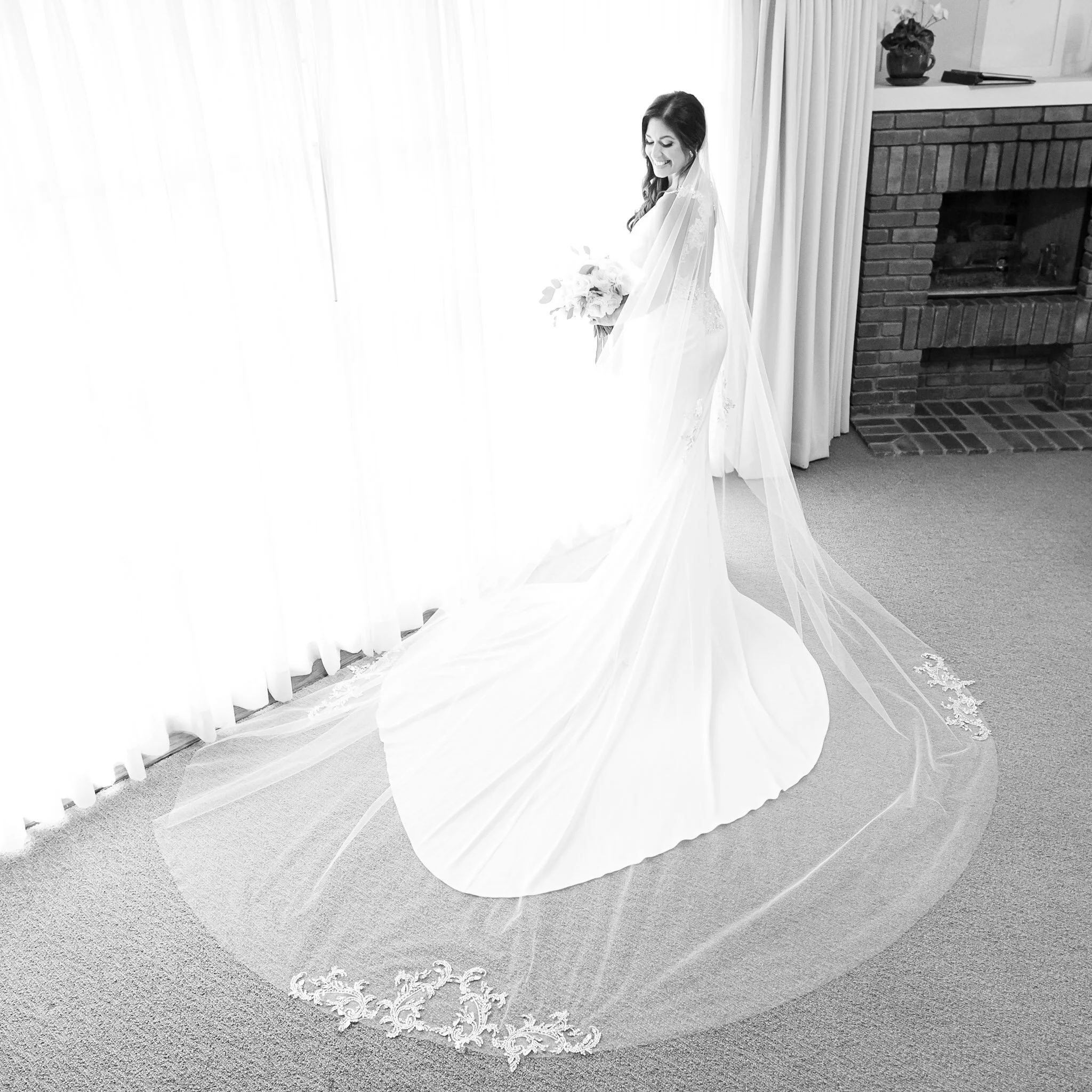 A woman in a wedding dress holding a bouquet, smiling, standing near a brick fireplace with a shelf, curtains, and carpeted floor.