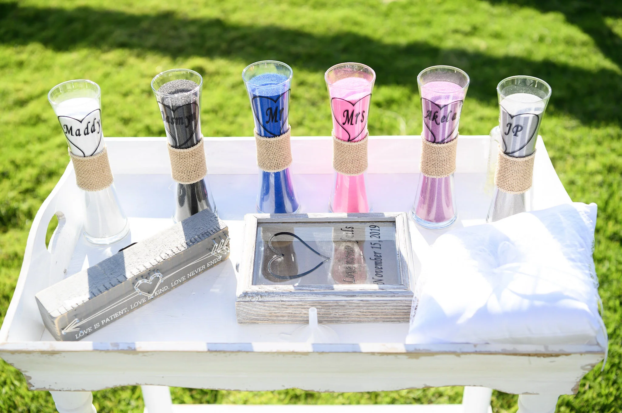 Six champagne flutes filled with sand in different colors, each labeled with a wedding title such as 'Maddie,' 'Groom,' 'Mr,' 'Mrs,' 'A Reba,' and 'J P,' placed on a white table outdoors with a grassy background.