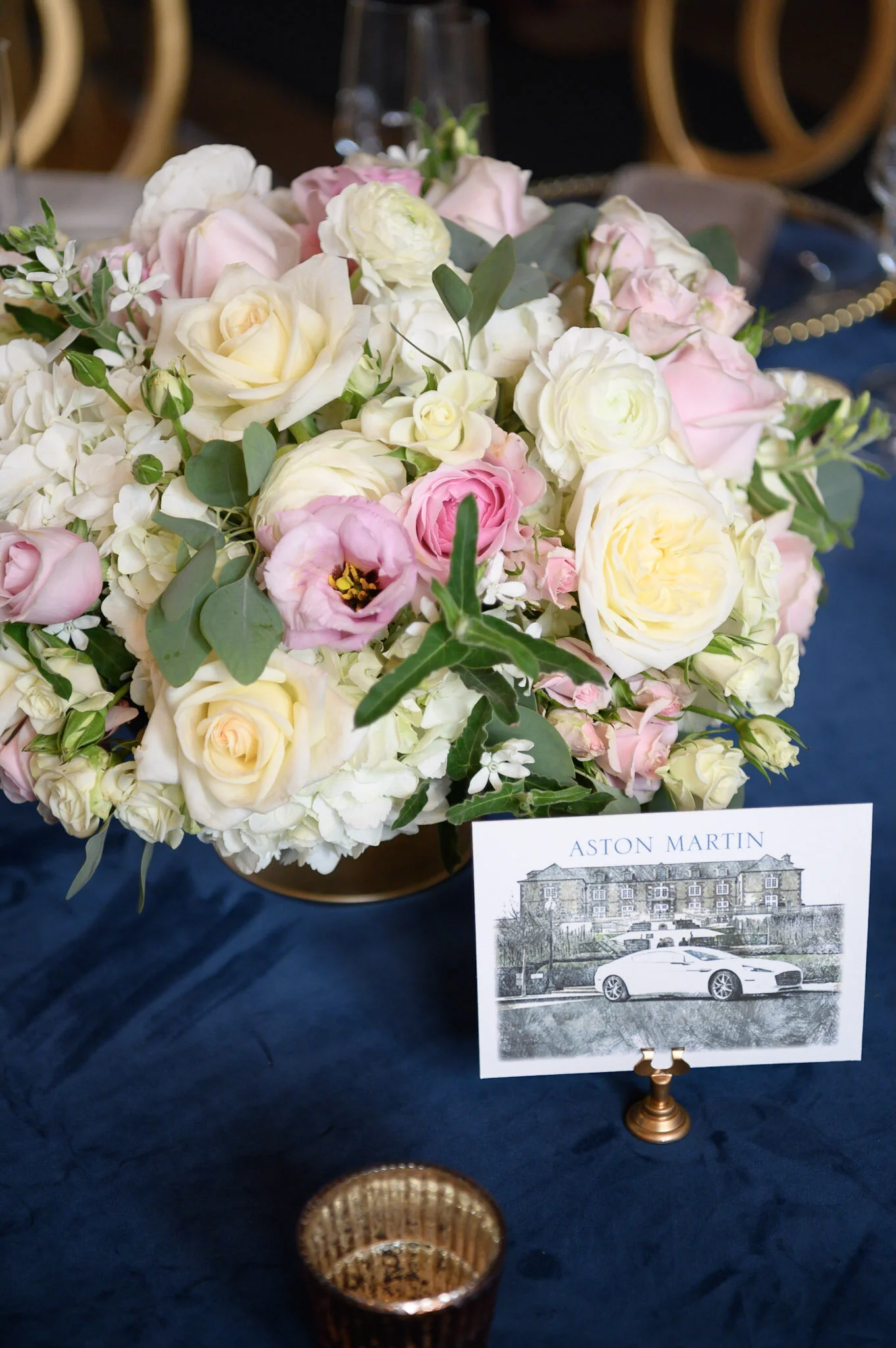 A floral arrangement with white, pink, and cream-colored roses and greenery on a dark table. A card with an illustration of a house and a car, labeled 'Aston Martin,' is placed next to the flowers.