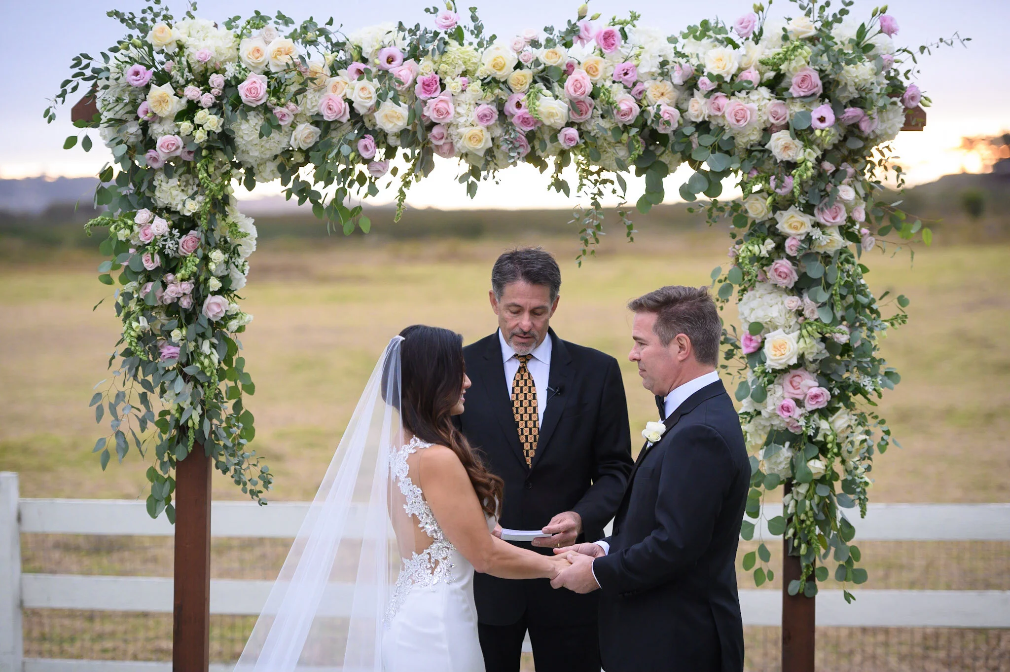 A wedding ceremony outdoors with a couple holding hands and facing each other, standing under a floral arch with pink and white roses and greenery, with an officiant officiating.