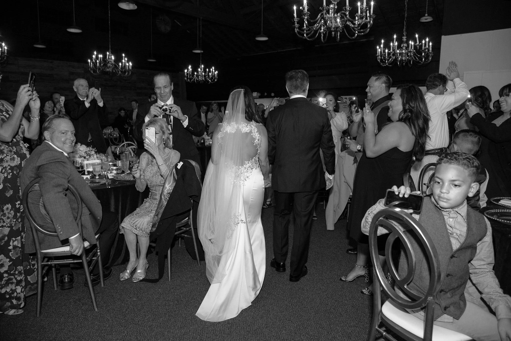 Wedding reception with bride in a veil and wedding gown, surrounded by guests taking photos and smiling in a dimly lit room with chandeliers.