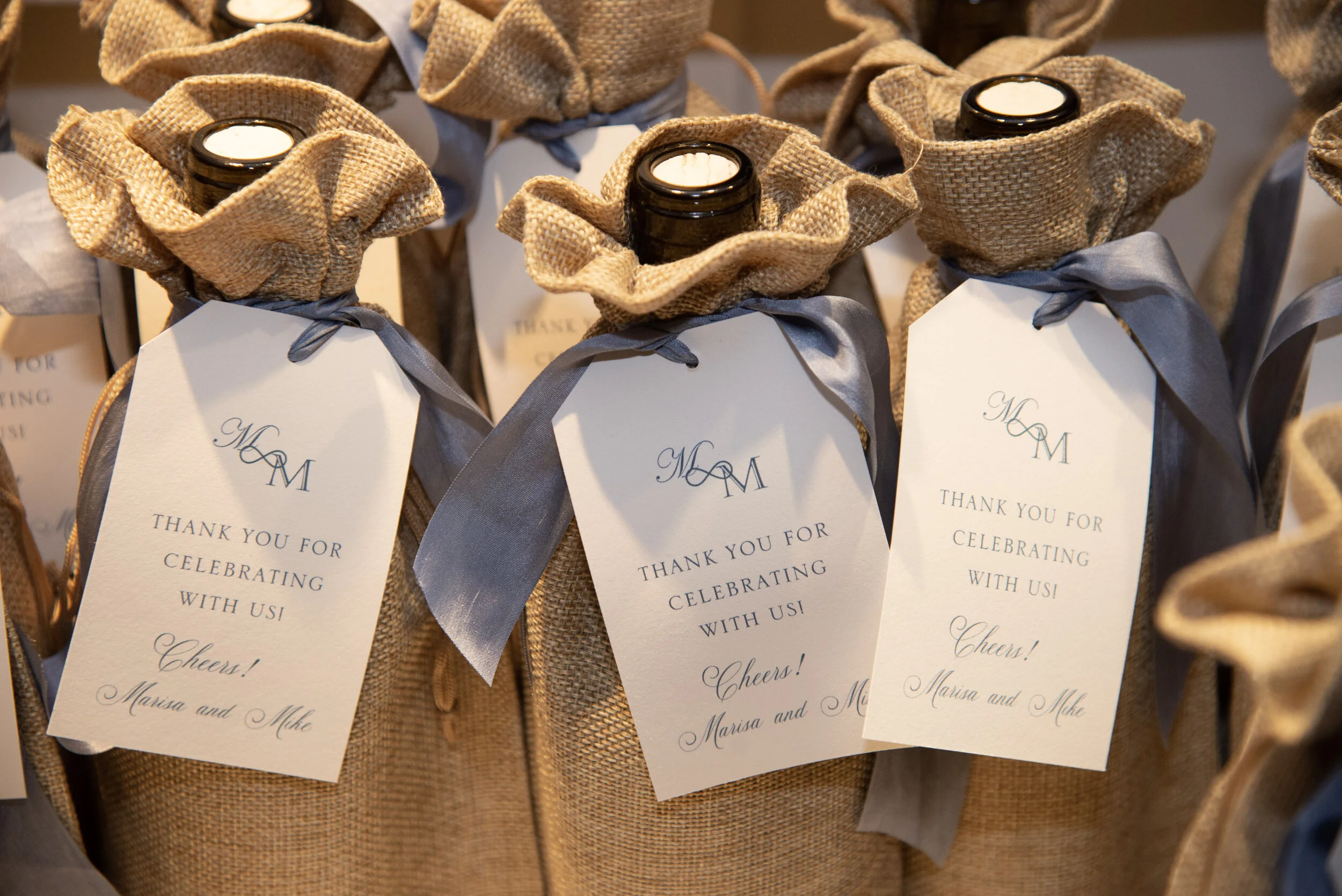 Bottles wrapped in burlap fabric with gray ribbons and thank you tags attached, likely for a celebration or event.