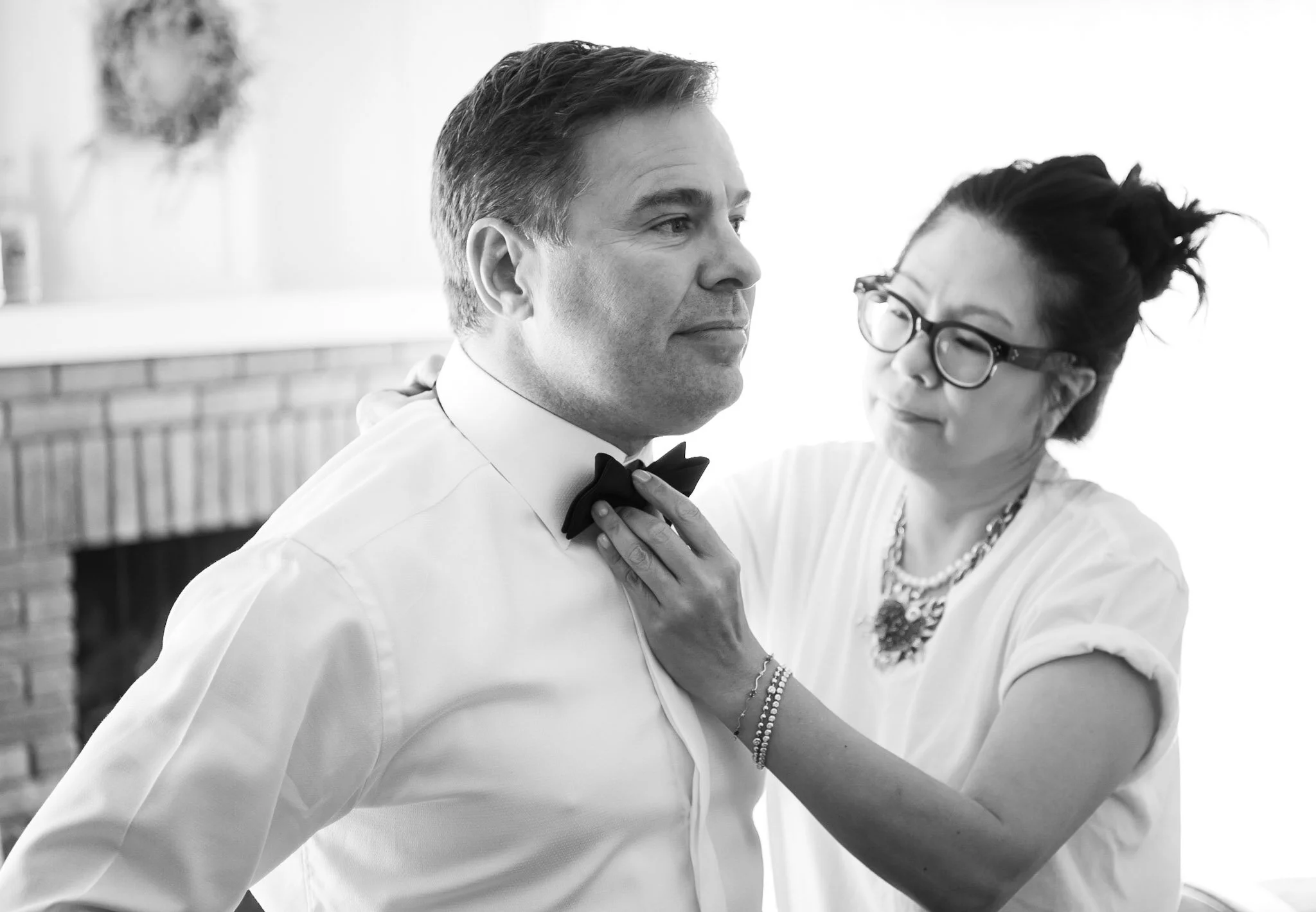 A man in a tuxedo and bow tie dancing with a woman who is wearing glasses and a white top, in a social setting with a brick wall in the background.