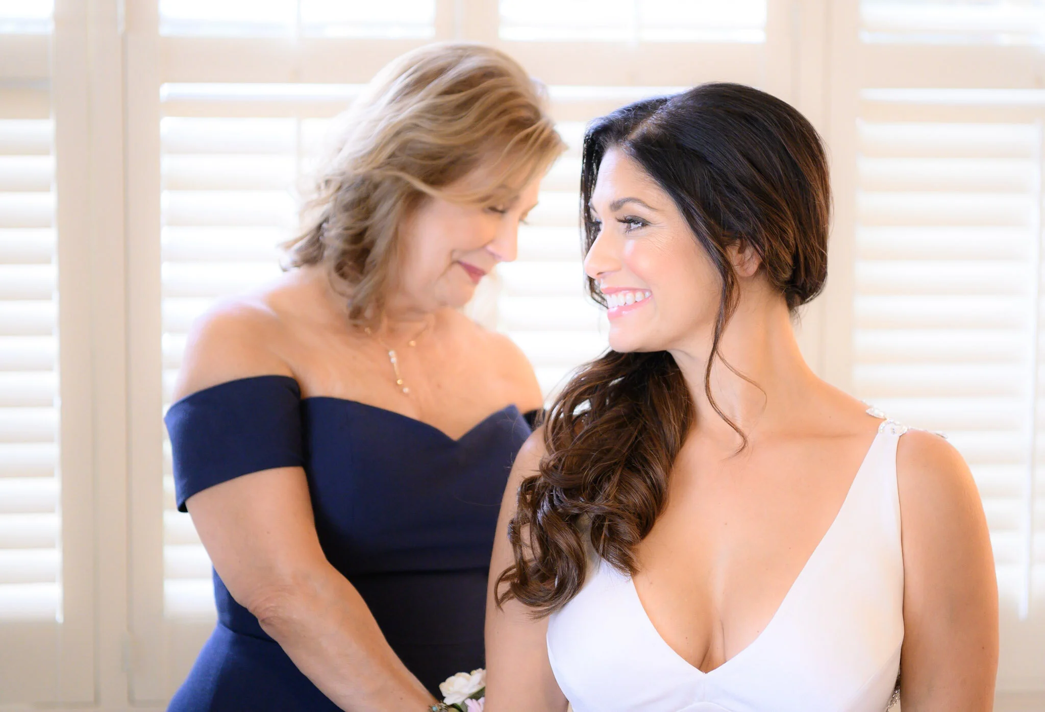 An older woman and a younger woman sharing an intimate moment, with foreheads touching and smiling warmly at each other, inside a room with white blinds.