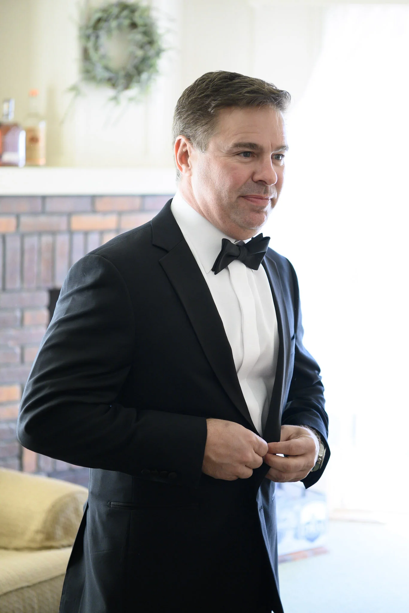 A man dressed in a black tuxedo with a bow tie, standing indoors near a yellow sofa with a brick wall and window behind him.