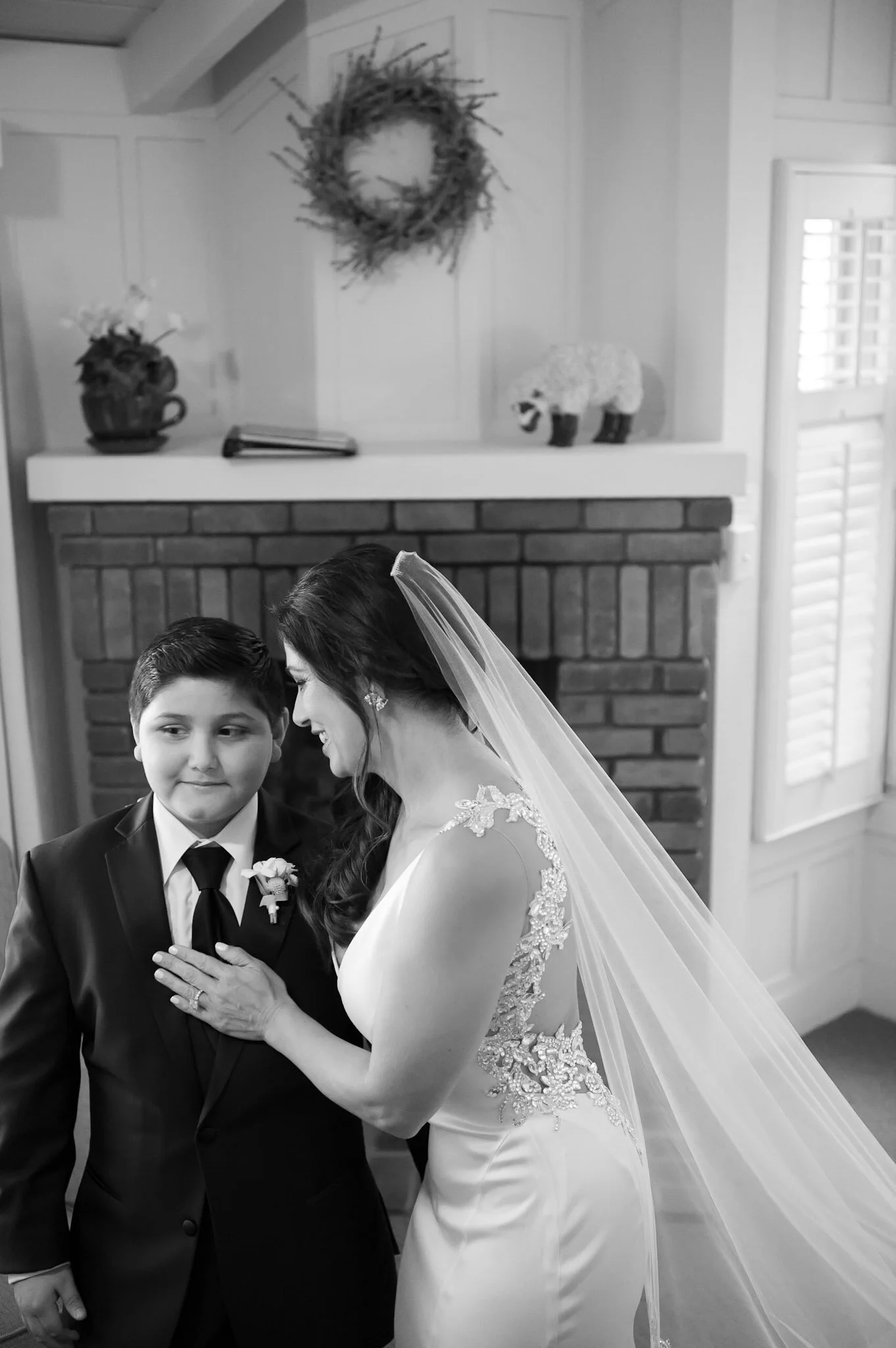 A bride and a young man are sharing a moment during a wedding ceremony indoors, with a brick fireplace and decorated mantel behind them.