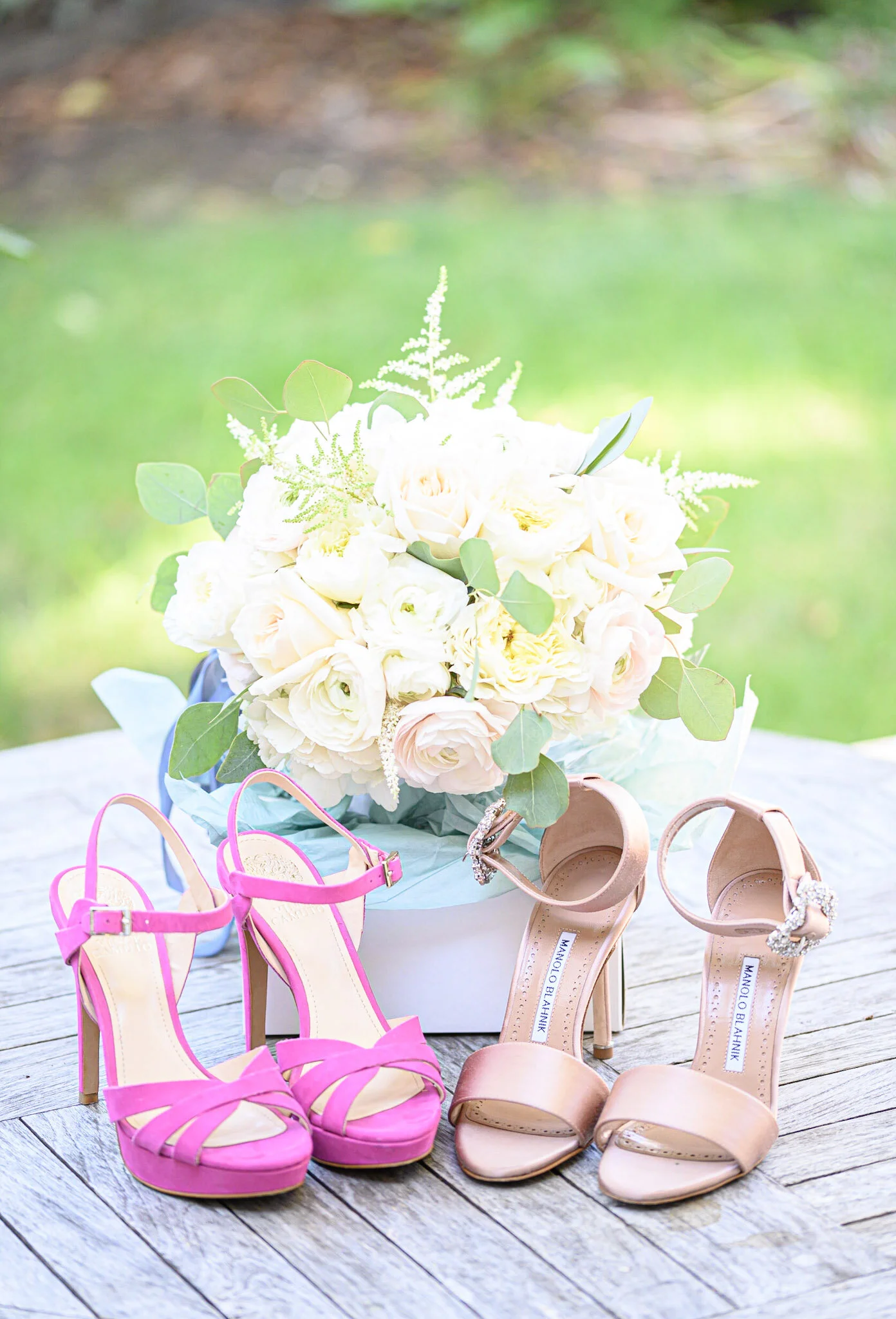 A bouquet of white and pink flowers with green leaves sits on a wooden surface, surrounded by four pairs of women's high-heeled shoes in pink and nude colors with decorative embellishments.