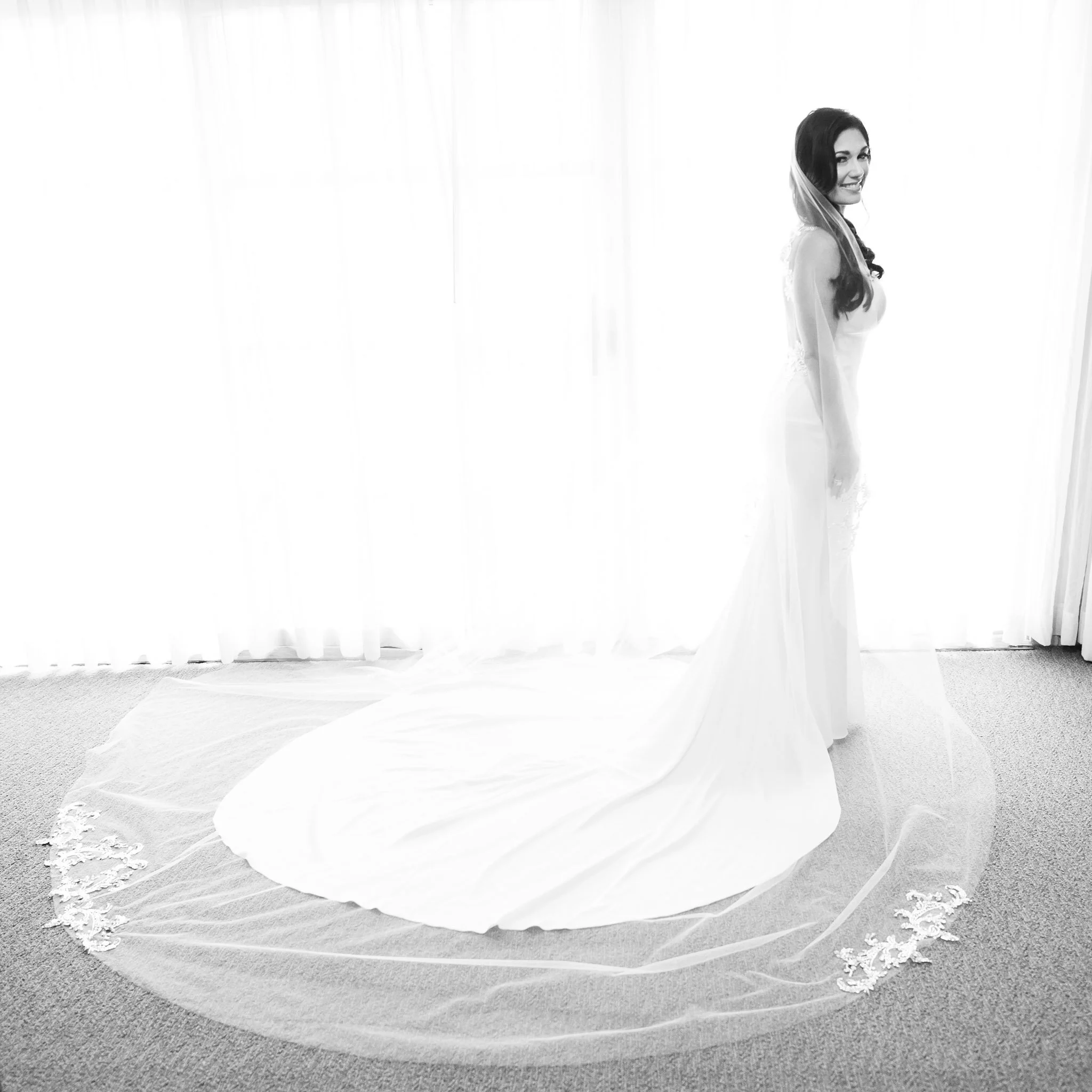A wedding dress is draped on a circular platform in front of a softly lit, sheer curtain, with a woman smiling and looking over her shoulder.
