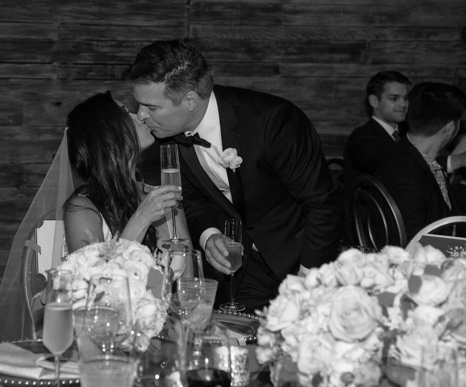 A wedding reception scene in black and white, featuring a bride and groom sharing a kiss at their table decorated with flowers and champagne glasses, with other guests in the background.