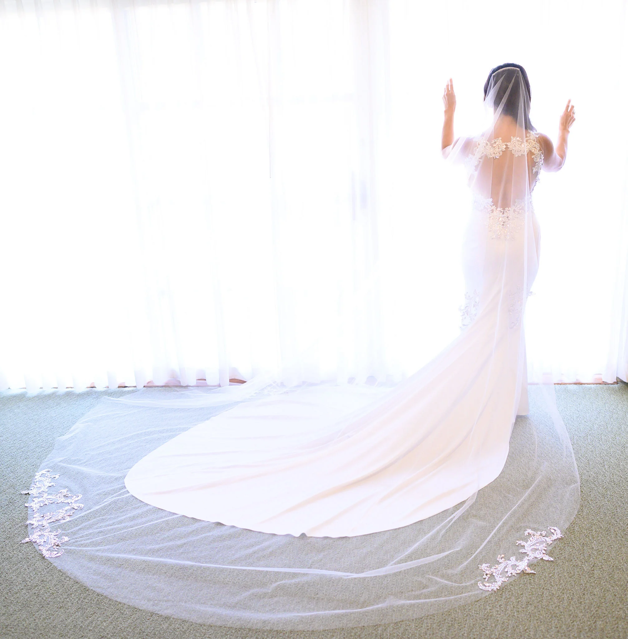 A bride in a wedding gown with lace detailing on the back, standing with her arms raised near a large window with white curtains, and a flowing veil over her dark hair.