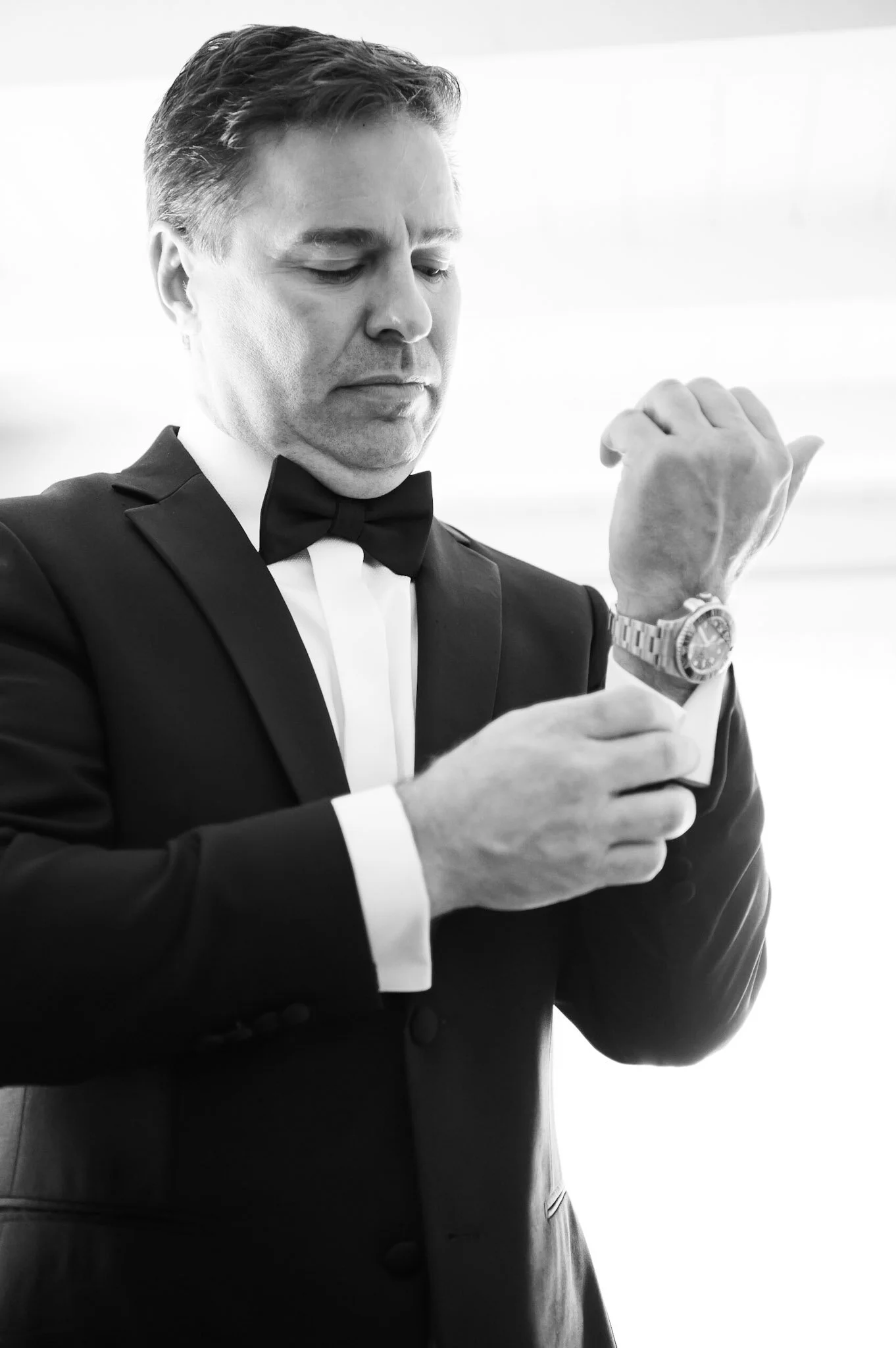 A man in a tuxedo with a bowtie, wearing a wristwatch, checking the time on his watch.