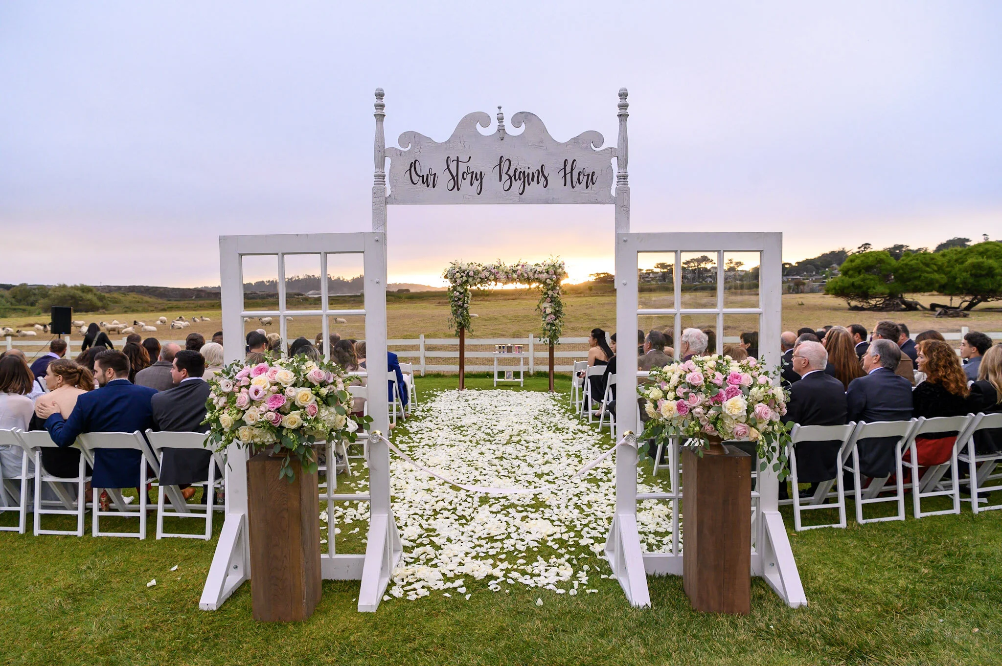 Outdoor wedding ceremony setup with white chairs, floral arrangements, an arch with a sign that says 'Our Story Begins Here,' and a scenic field at sunset.