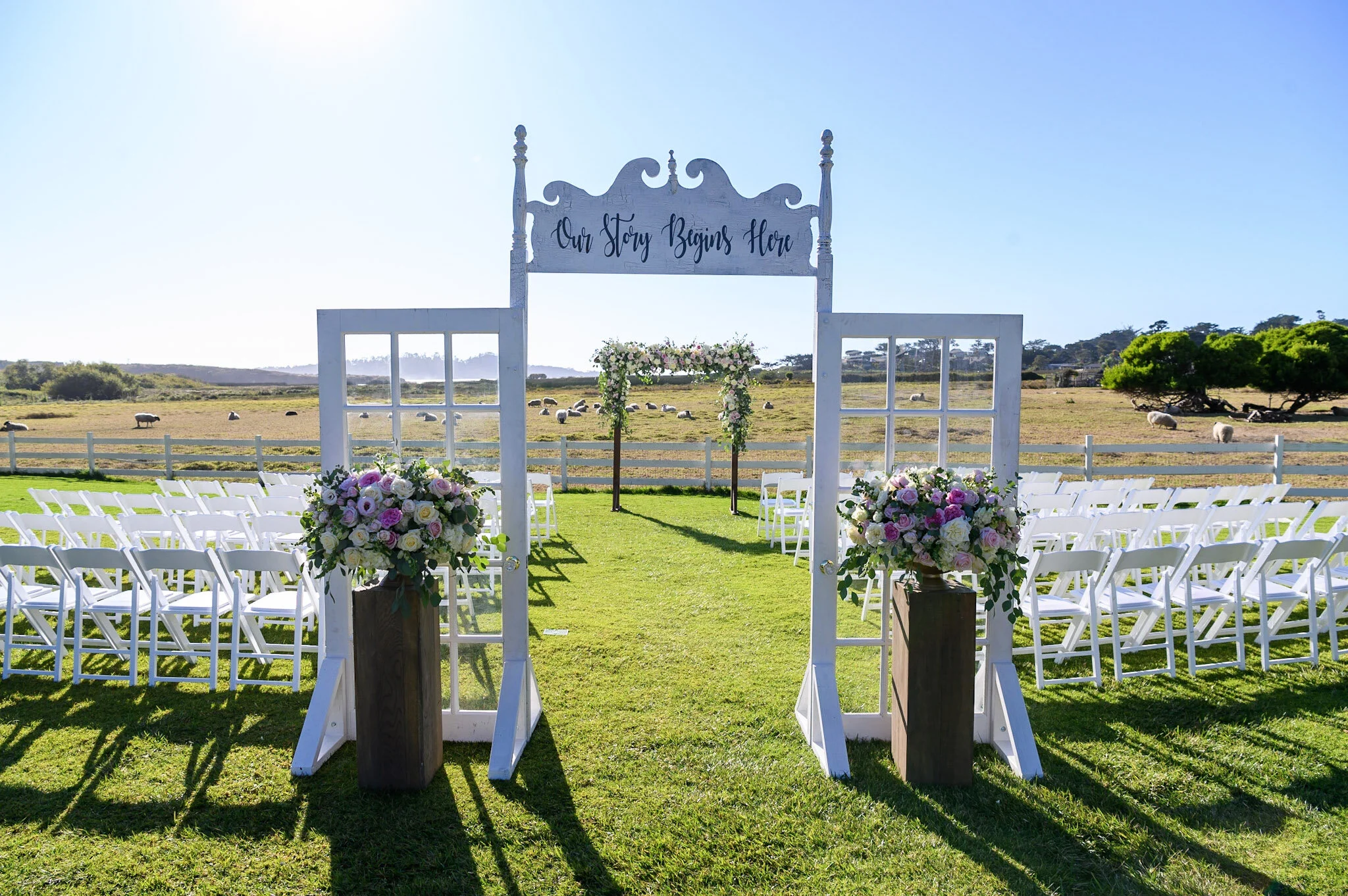 Outdoor wedding setup with white chairs arranged in rows on green grass, floral arrangements, a white arch with a sign that says 'Our Story Begins Here,' and a scenic background of a field with grazing sheep and trees under a clear blue sky.