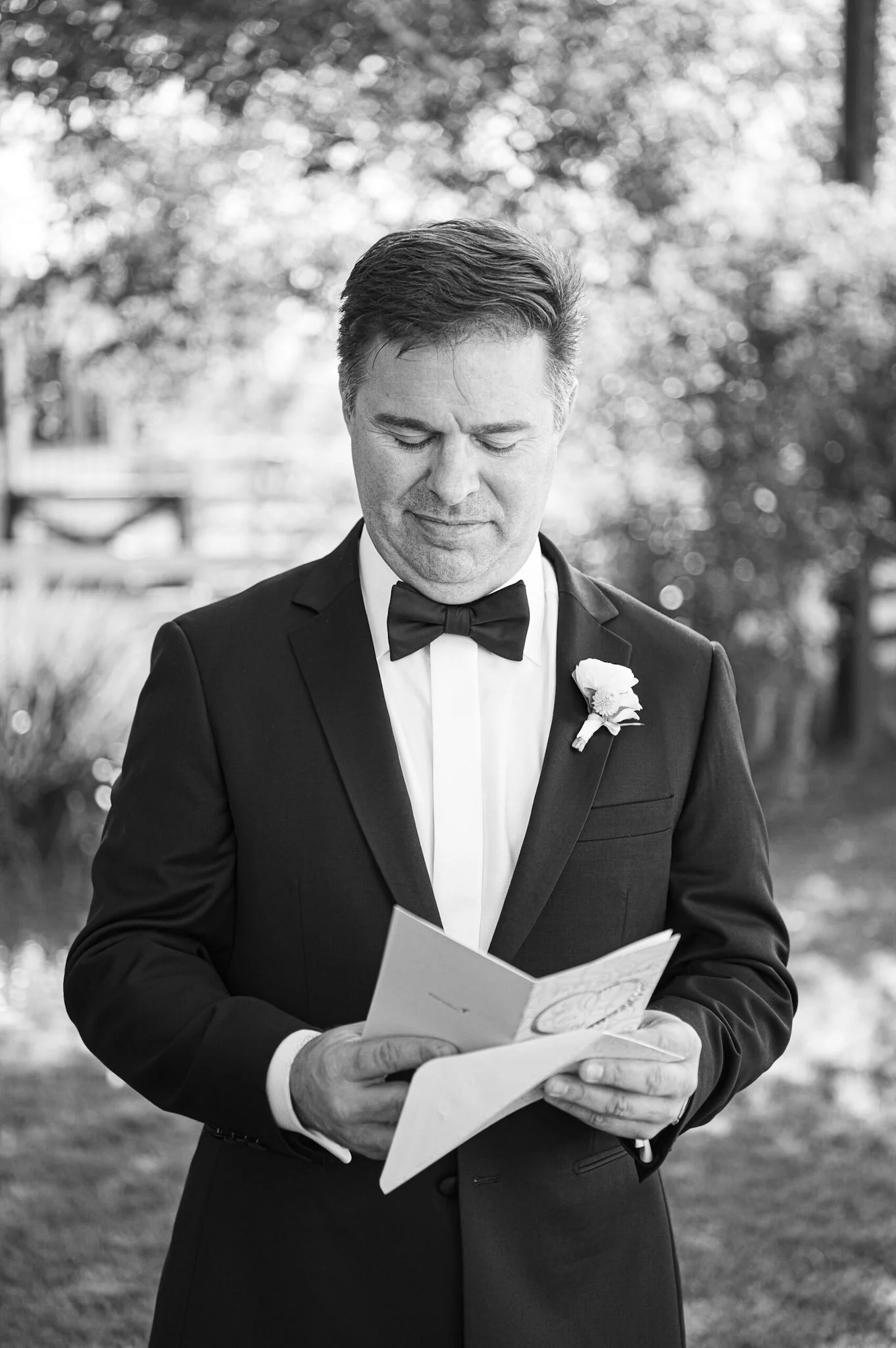 A man in tuxedo reading a card outdoors with trees in the background.