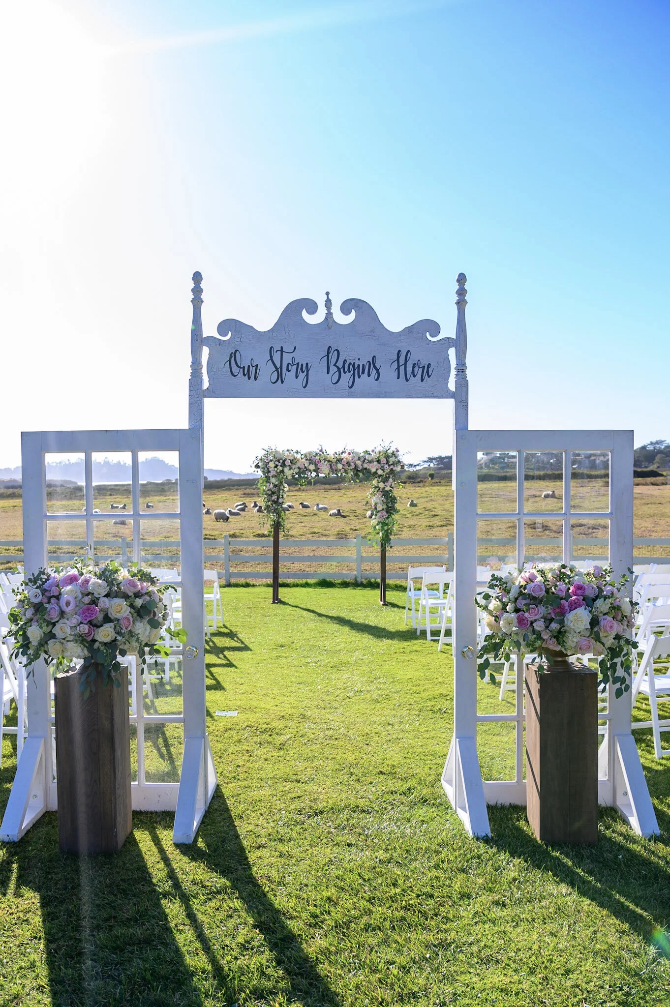 Wedding ceremony setup outdoors with white chairs, floral arrangements, and a wooden arch with a sign that reads "Our Story Begins Here" against a field and blue sky.
