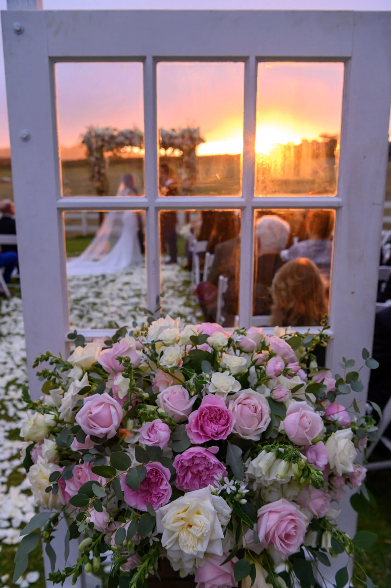 A floral arrangement of pink, white, and cream roses with green foliage, placed in front of a window showing a sunset scene with trees and people, during a wedding ceremony.