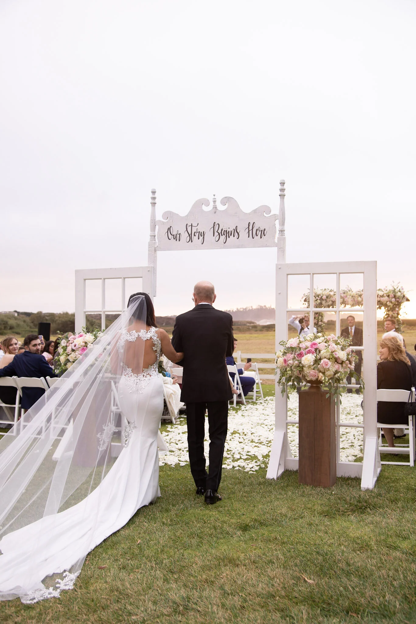 Bride and groom standing at the altar during an outdoor wedding ceremony. The bride wears a white wedding dress with a long train and veil, and the groom wears a black suit. The altar has a sign that reads "Our Story Begins Here" and is decorated wit