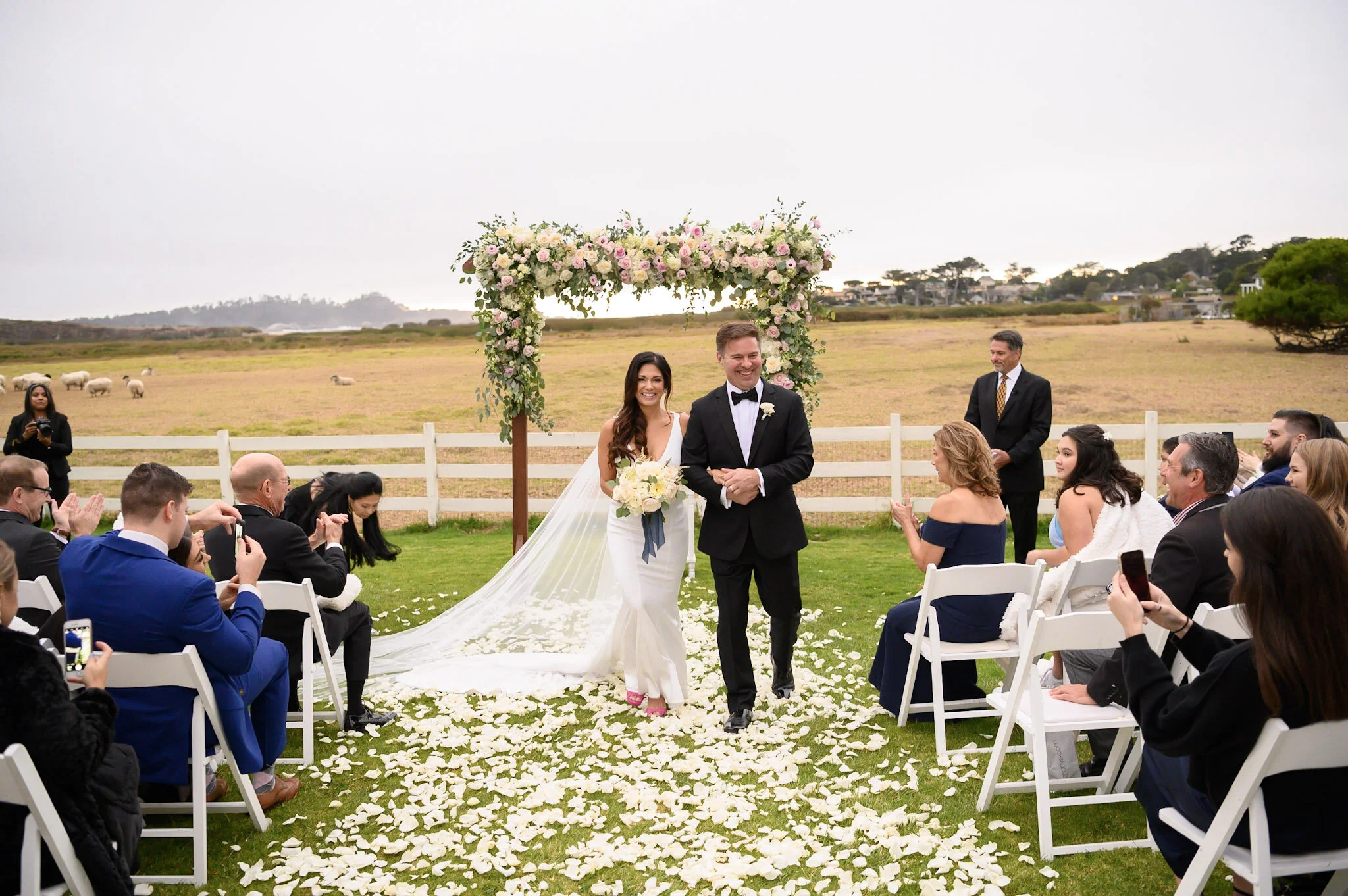 A bride and groom walking down the aisle at an outdoor wedding ceremony with guests clapping and taking photos, floral arch, green field, cloudy sky.