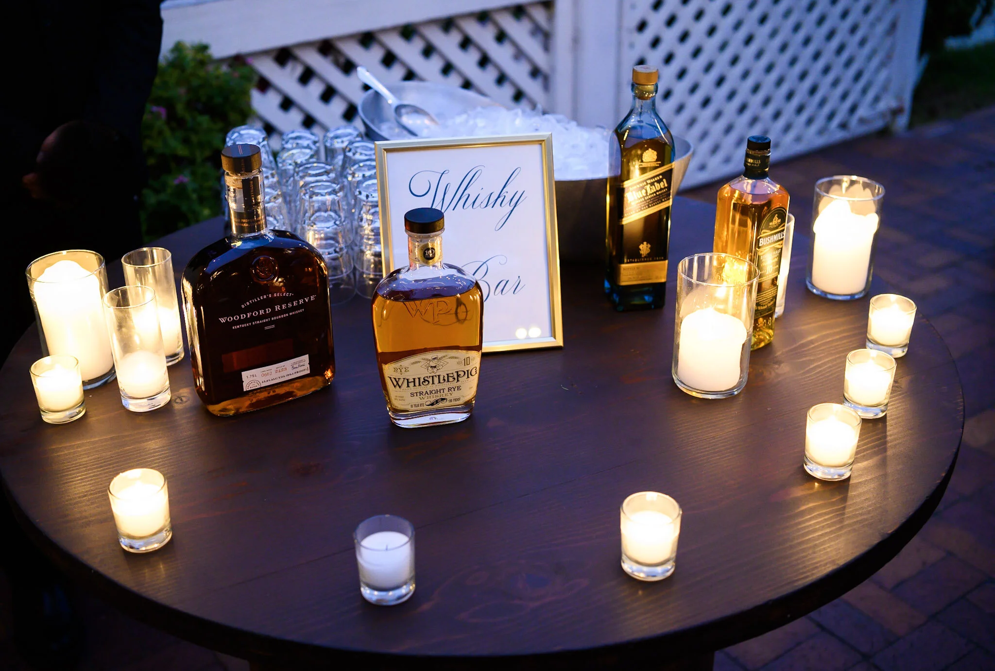 Table with bottles of whiskey, glasses, a small framed sign reading 'Whisky Bar', and numerous candles in glass holders, set outdoors with a lattice fence in the background.
