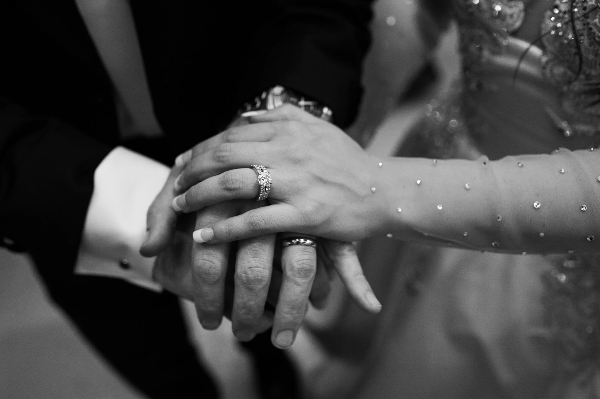Close-up of a bride and groom holding hands, showing wedding rings, with the bride's hand resting on top of the groom's hand.