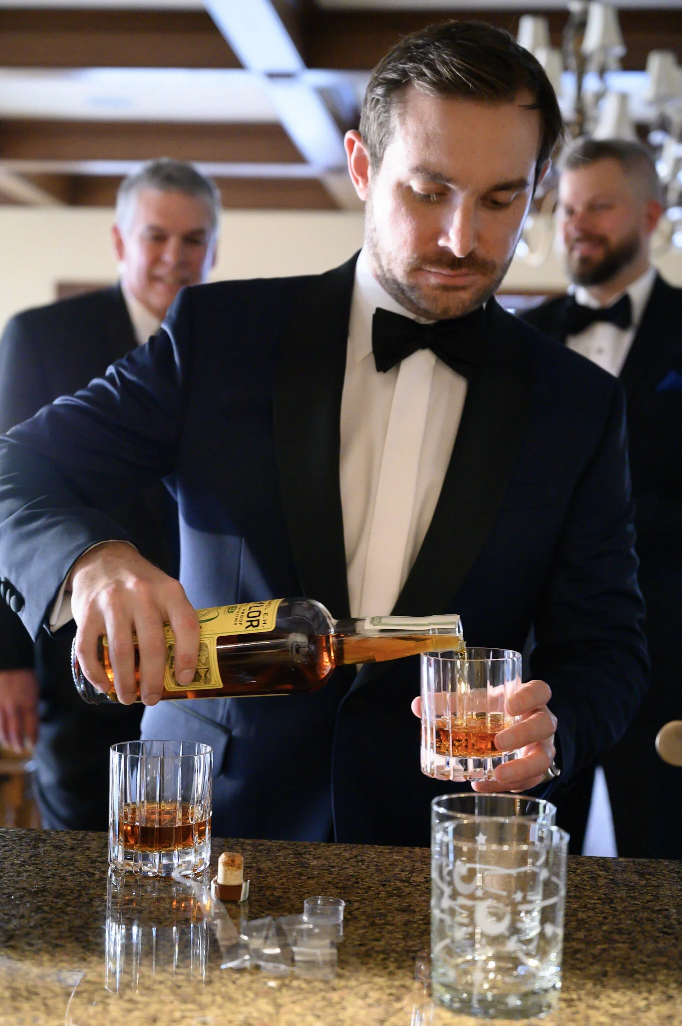 A man in a tuxedo pouring whiskey into a glass at a bar, with two other men in tuxedos in the background, and several empty glasses on the bar counter.