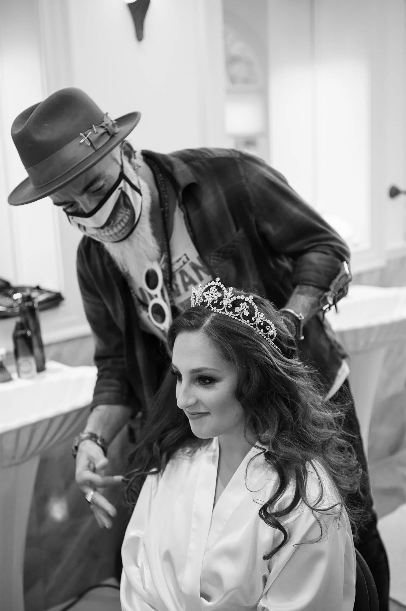 A woman with long, wavy hair wearing a crown sits while a man wearing a wide-brimmed hat, glasses, and a mask leans over her, smiling in a black-and-white photo.