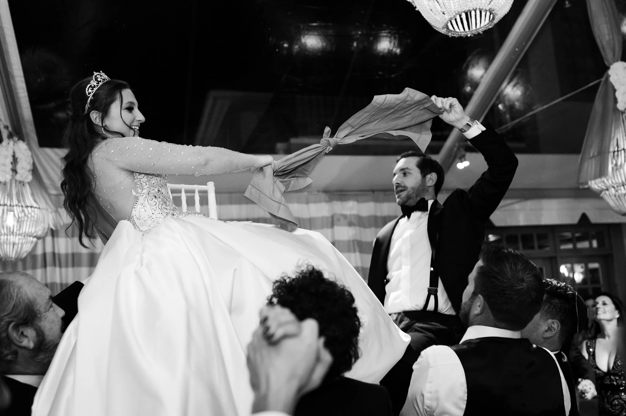 A woman wearing a tiara and a wedding dress is being lifted by a man in a tuxedo while they dance at a wedding reception. The woman is smiling and holding a cloth, and there are guests watching in the background.