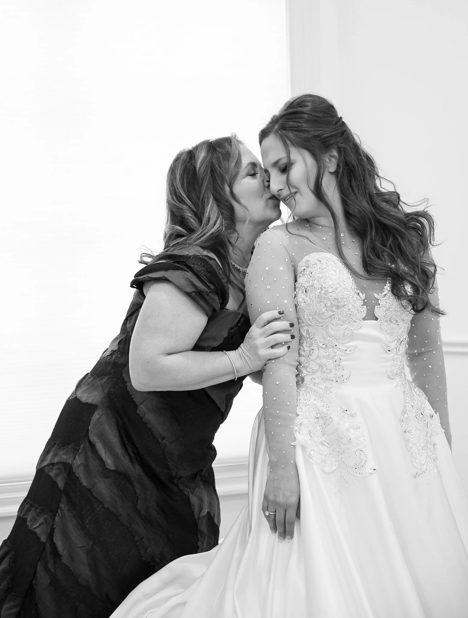 A woman in a black dress leans in to kiss a woman in a wedding gown on the cheek. They are smiling and holding hands, with the woman in the wedding dress clutching her gown.