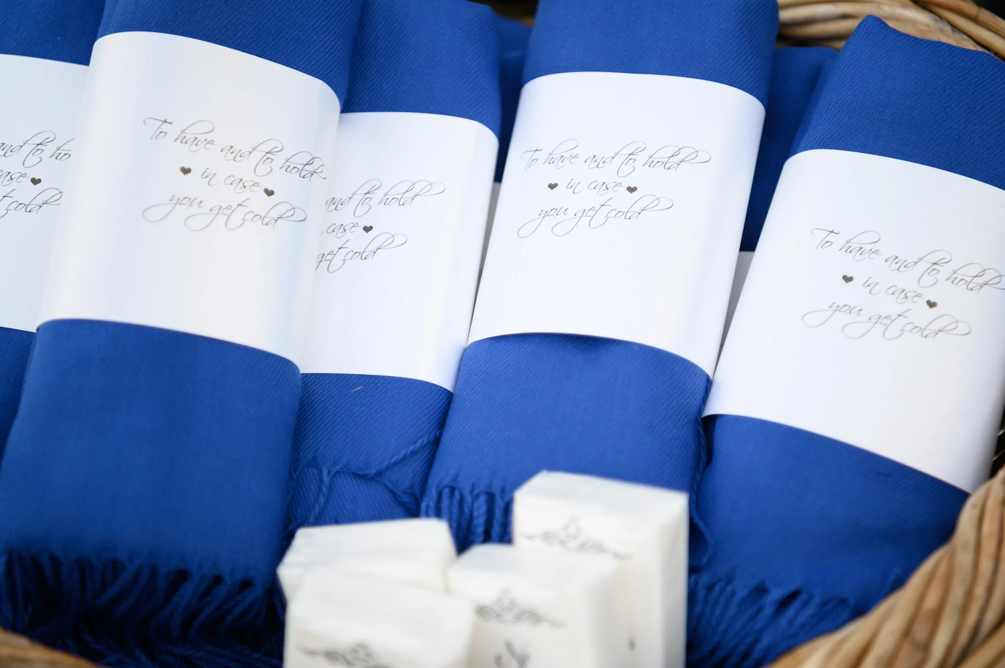 Close-up of white paper napkins in a basket, with a white phrase and small heart icons on each napkin that reads: 'To have and to hold in case you get cold.'