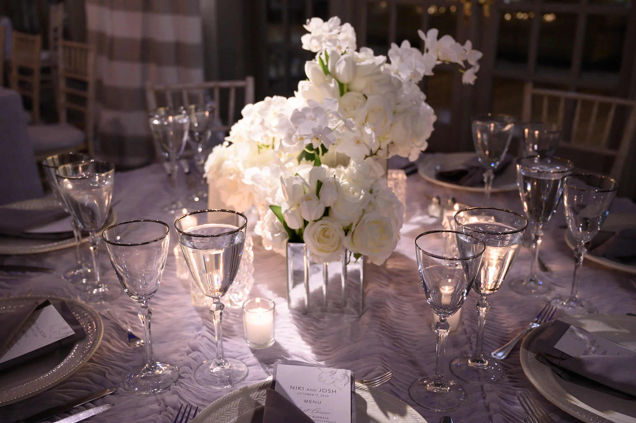 A dinner table decorated with a tall floral centerpiece of white roses and orchids, surrounded by wine glasses, silverware, and place settings, with dim lighting and chairs in the background.