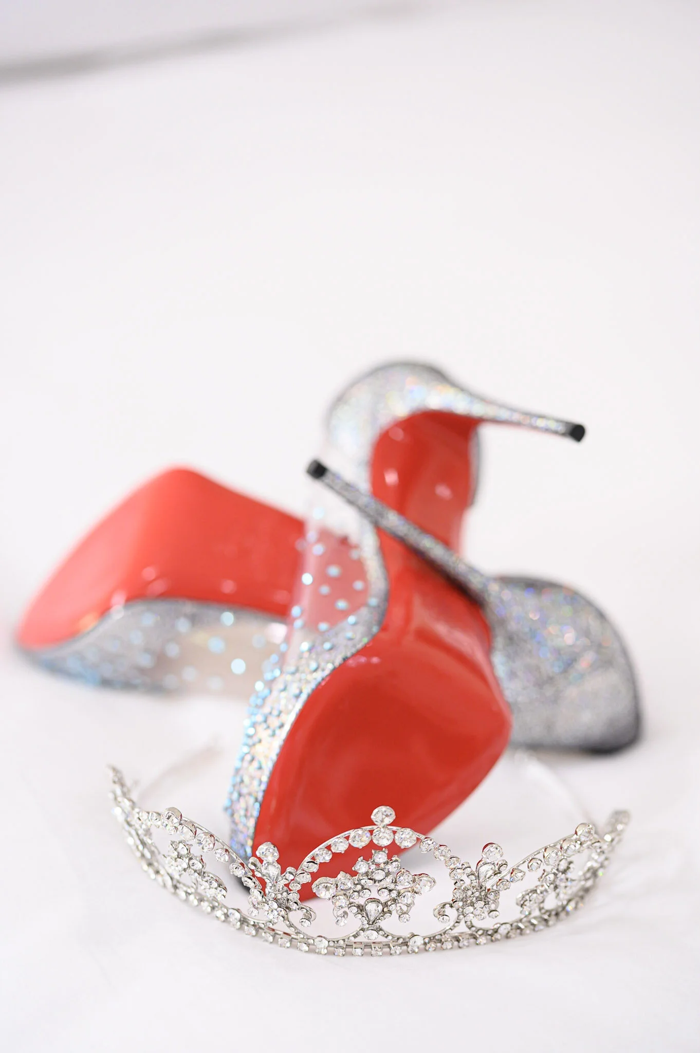 A silver tiara decorated with clear rhinestones resting on a white surface. Behind it, a red high heel shoe with a glittery main strap and a thin black heel is visible.
