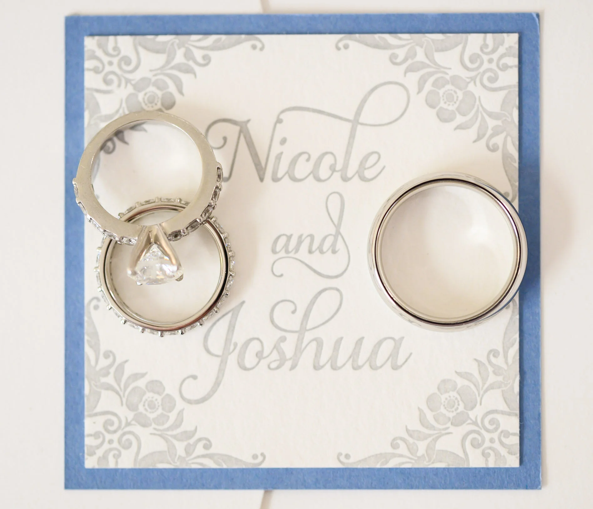 A greeting card with silver lettering that says "Nicole and Joshua" on a decorative floral background, with two silver rings placed on top.