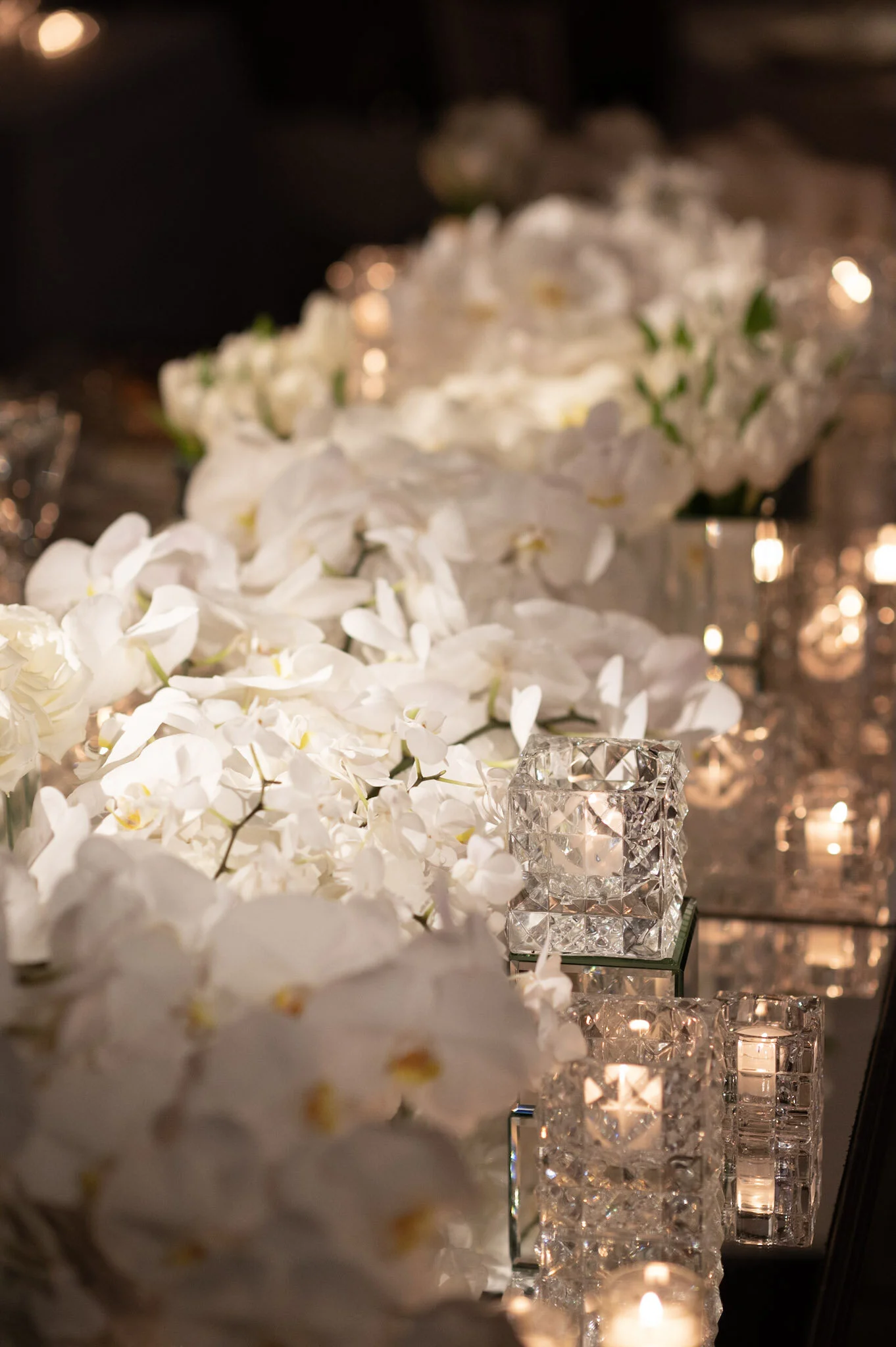 Close-up of white orchids and candles in glass holders on a reflective surface, with blurred background of candlelight reflections.