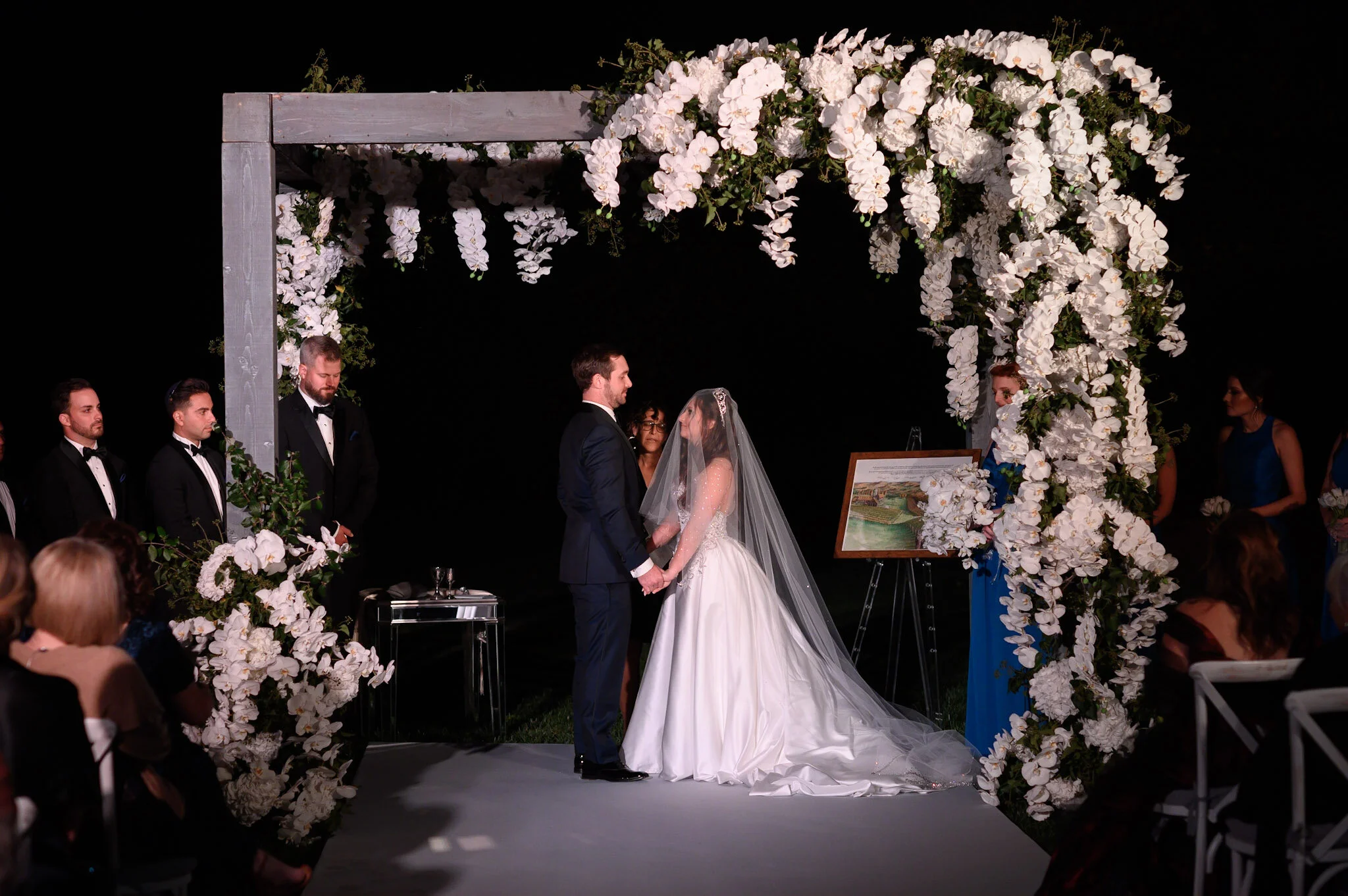 A wedding ceremony with a bride and groom holding hands under a floral arch at night.