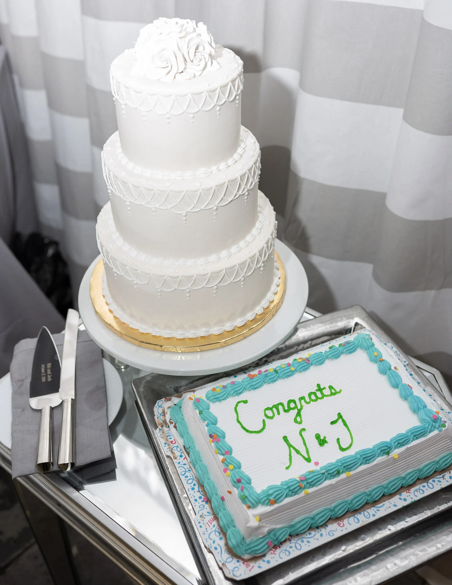 A three-tiered white wedding cake with piped decorations on each tier, topped with roses, and a rectangular sheet cake with blue piping and green writing, on a silver tray at a celebration event.