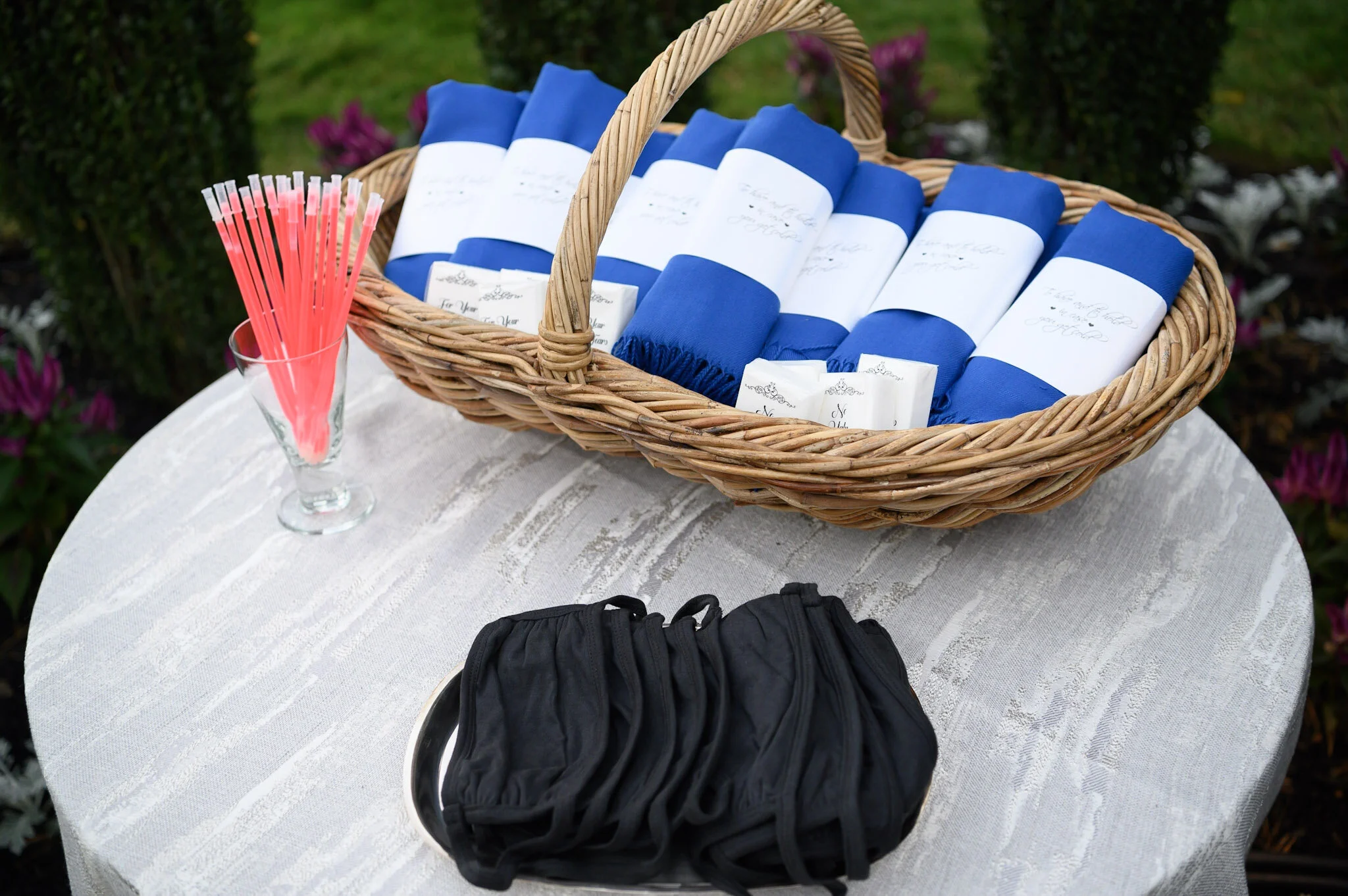 A wicker basket with blue and white rolled fabric items, a glass filled with pink drink straws, and a pair of black gloves on a white lace tablecloth outside, with greenery in the background.