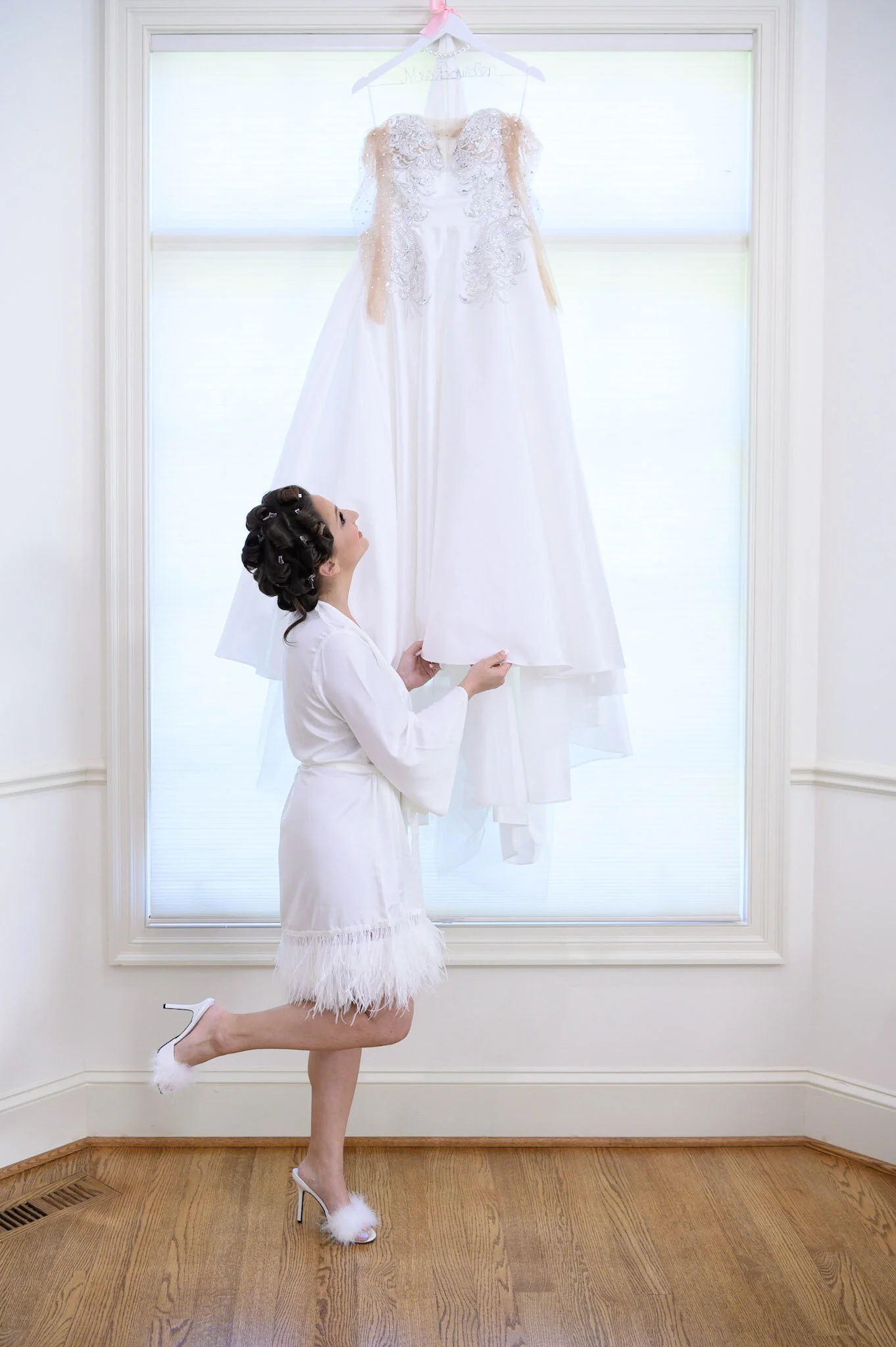 A woman kneeling on one knee with her hair in curlers, wearing a white feathered robe and high heels, looking at a wedding dress hanging in front of a window.
