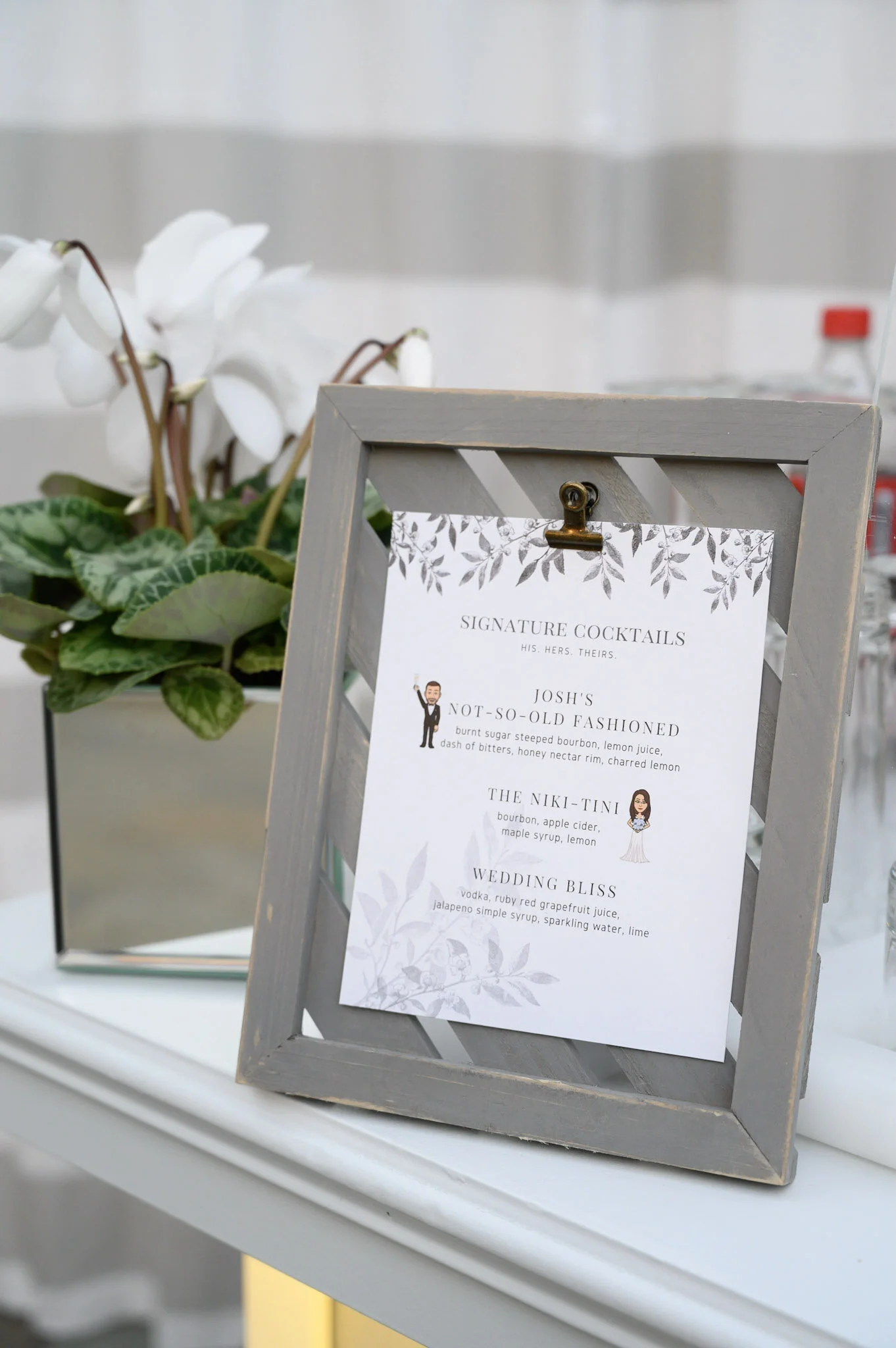 A decorative menu sign on a wooden frame displays cocktail names and ingredients, placed next to a pot of white orchids.