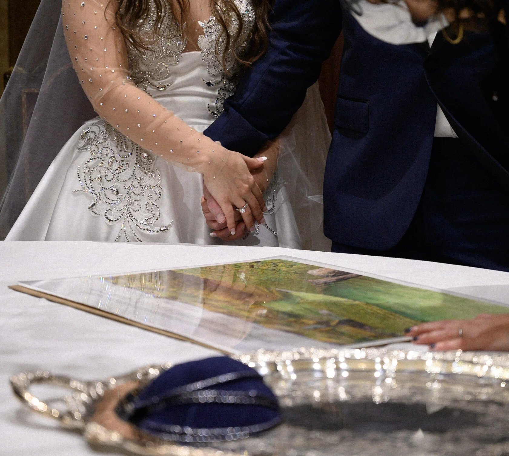 A bride and groom holding hands during a wedding ceremony. The bride is wearing a white dress decorated with rhinestones and lace, and the groom in a dark suit. There is a book or photo album on the table in front of them.
