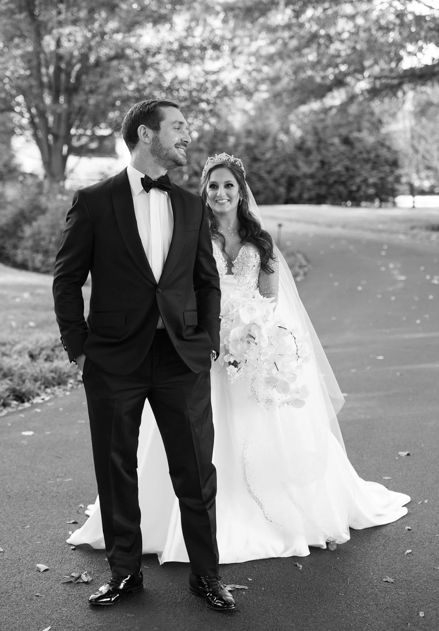 Black and white photo of a groom and bride on their wedding day outdoors. The groom is wearing a tuxedo with a bow tie, smiling while looking to his left. The bride is in a wedding gown with intricate beadwork, wearing a tiara, and holding a bouquet 