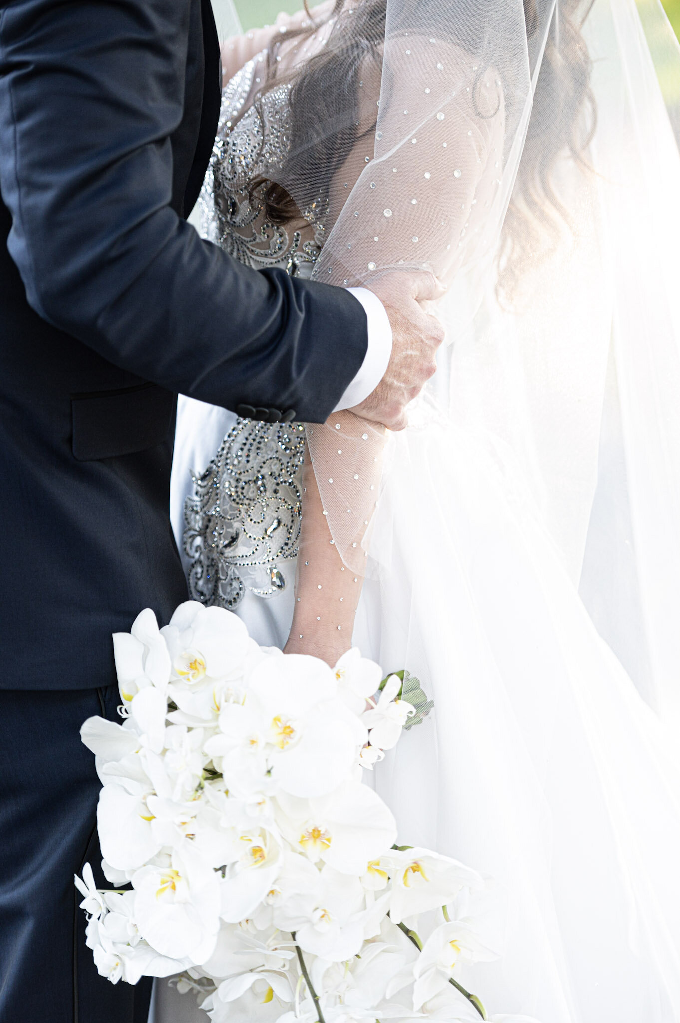 A bride and groom during a wedding ceremony, with the groom in a black suit and the bride in a white wedding dress with intricate beading and embellishments. The bride holds a bouquet of white orchids.