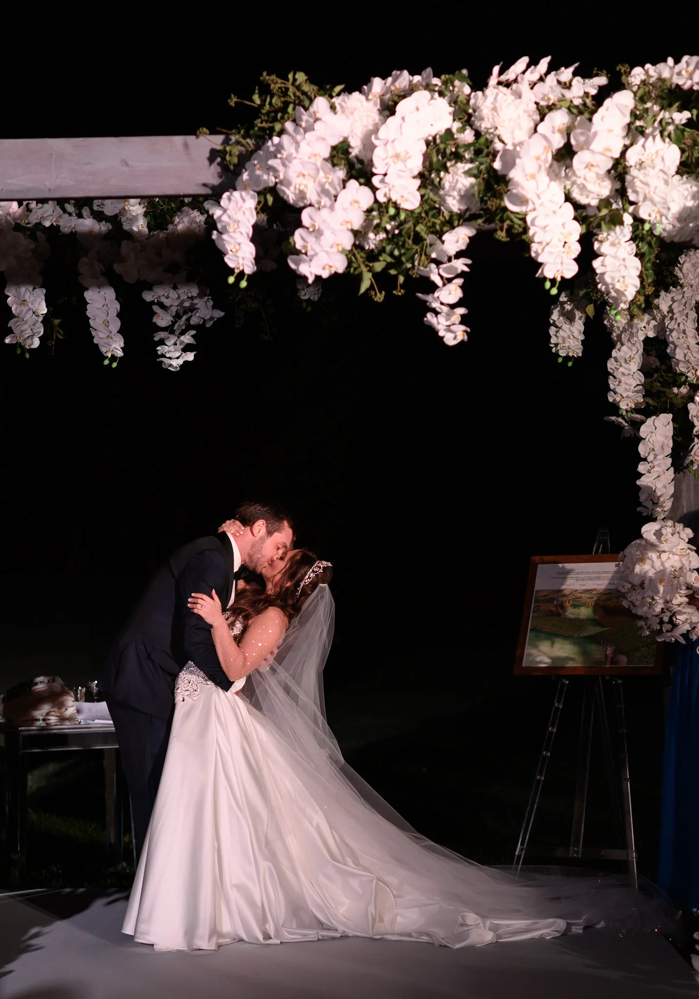 A bride and groom sharing a kiss during their wedding ceremony under a floral arch at night.