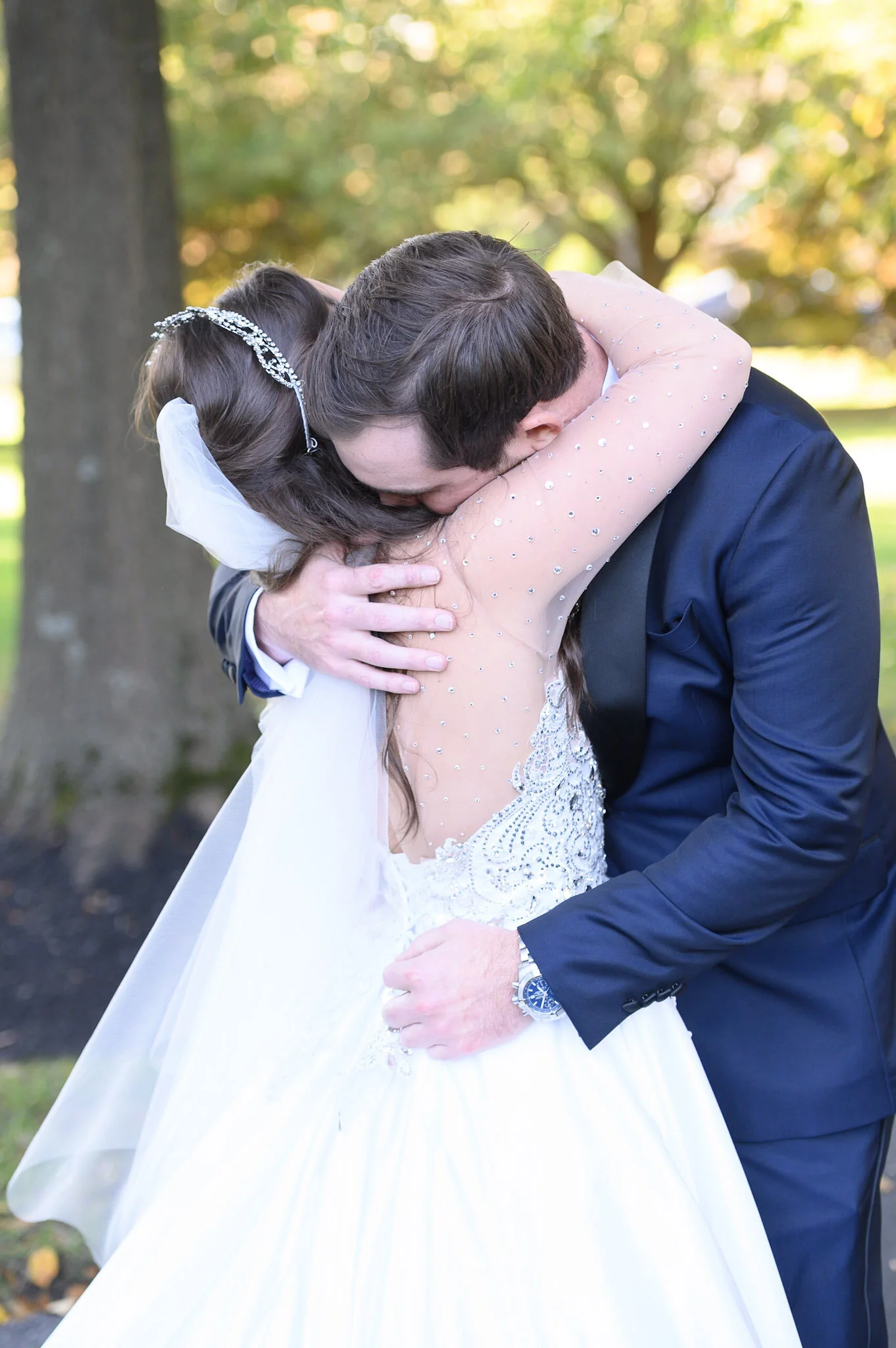 A bride and groom embrace outdoors, with trees and sunlight in the background, during their wedding celebration.