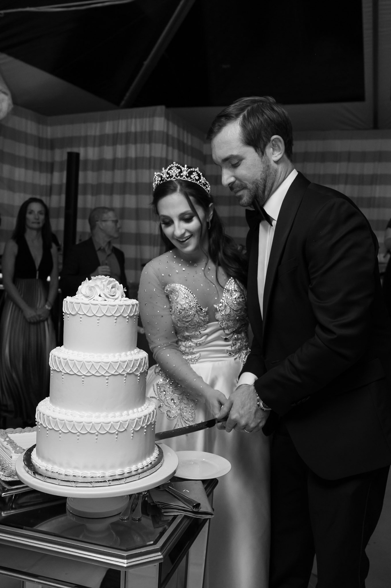 Wedding couple cutting a multi-tiered wedding cake together, with onlookers in the background, in a formal setting.