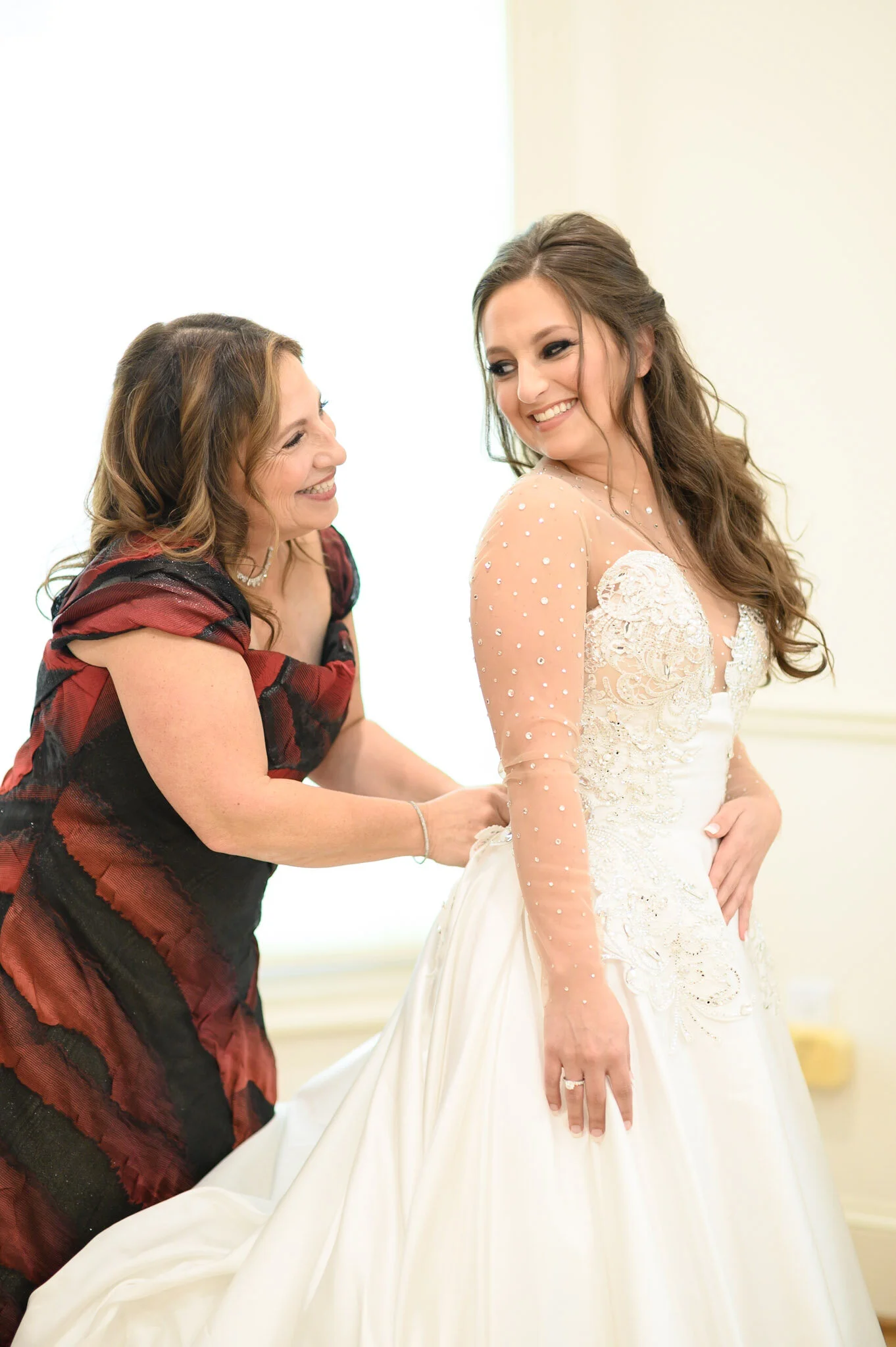 A woman in a wedding dress standing in front of another woman who appears to be helping her get ready for her wedding.