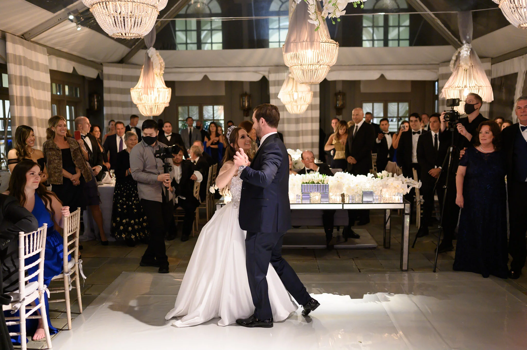 A bride and groom dance together in the center of a wedding reception hall filled with guests. The bride wears a white wedding gown and tiara, and the groom is dressed in a dark suit. The hall has large windows, elegant chandeliers, and floral decora