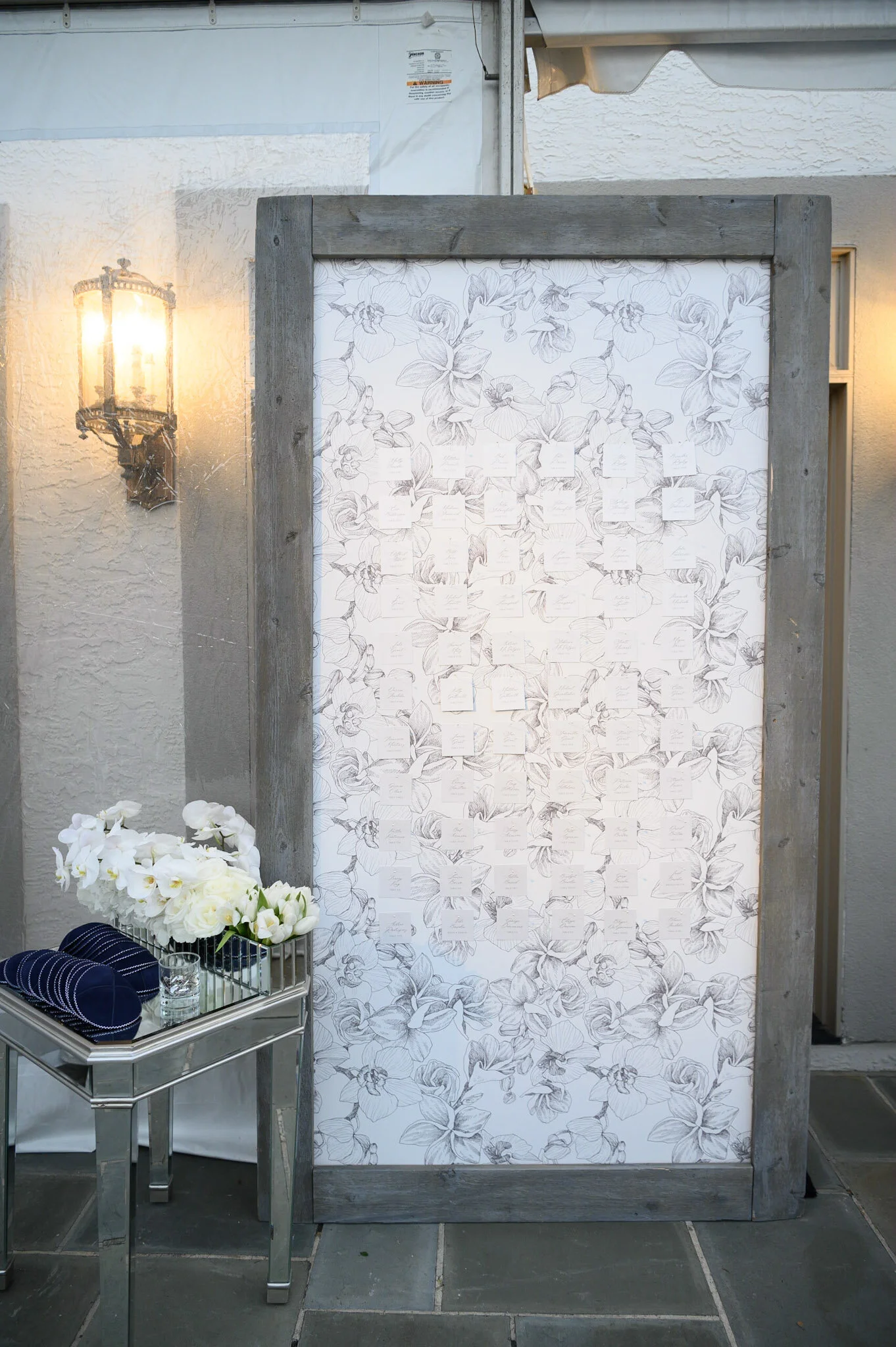 Decorative white floral panel with handwritten notes, flanked by an ornate wall-mounted lantern light, and a small table holding white flowers, glasses, and blue-striped items.