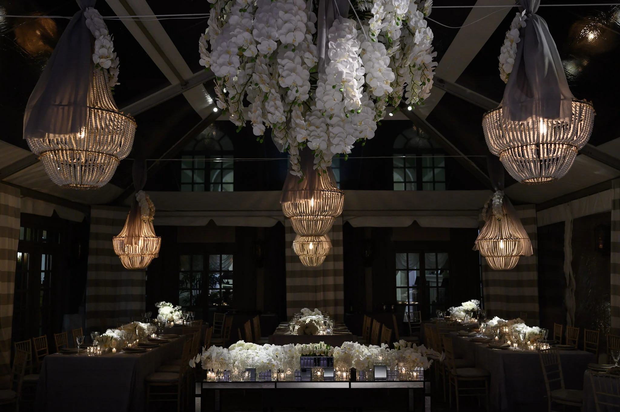 Elegant event space decorated with large white flowers, chandeliers with fabric and bead embellishments, and table settings with white flowers and candles.