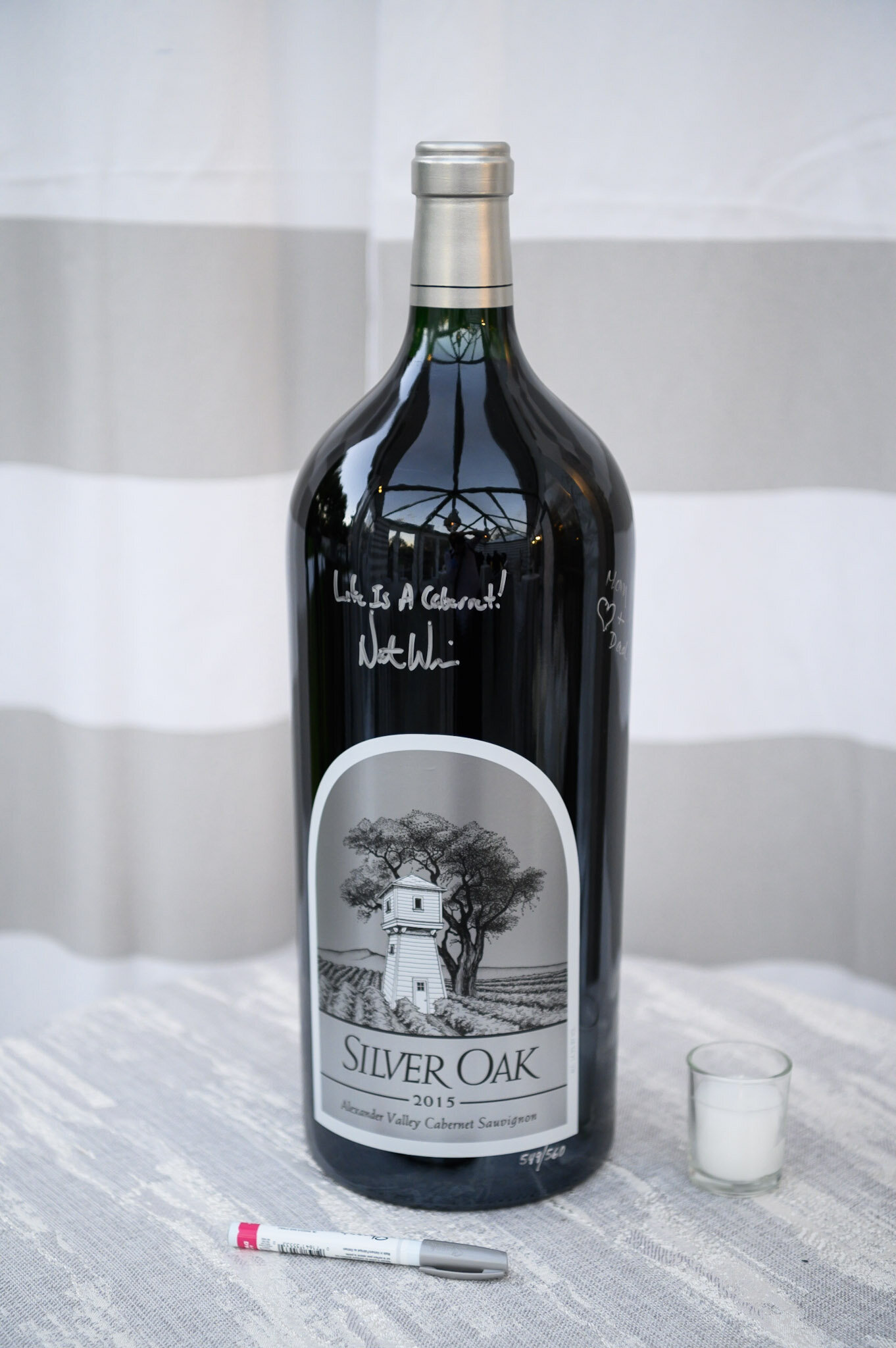 A bottle of Silver Oak Cabernet Sauvignon wine from 2015 with a black and white label featuring a tree and a house, placed on a table with a small white candle in a glass holder nearby, and a white marker pen in front of the bottle.