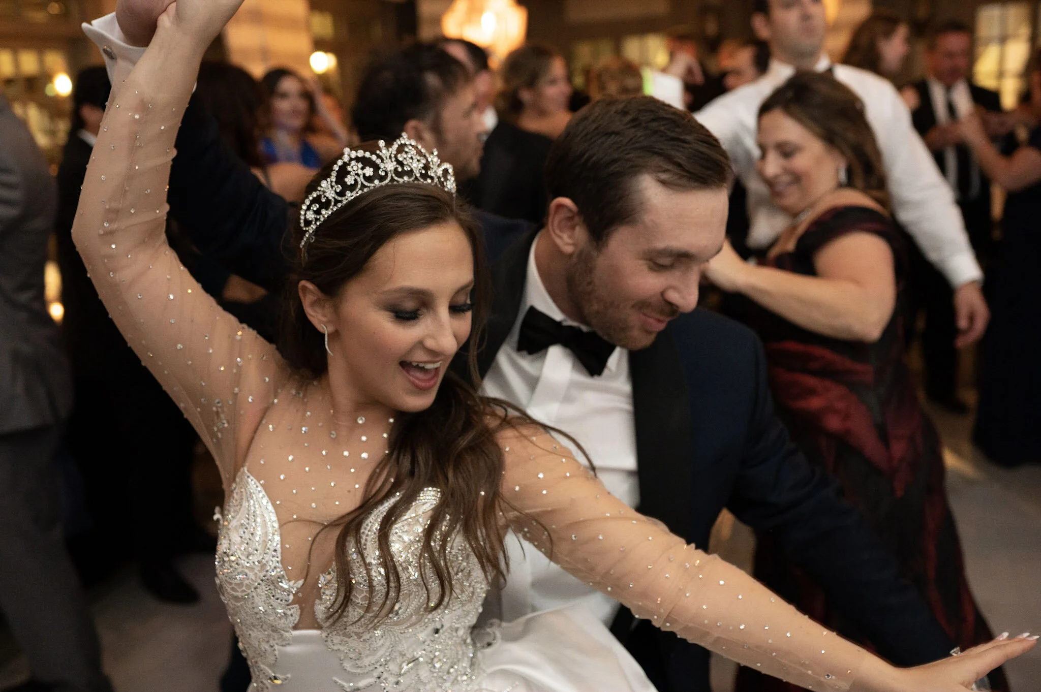 A bride and groom dancing at their wedding reception, surrounded by wedding guests in a warmly lit indoor venue.